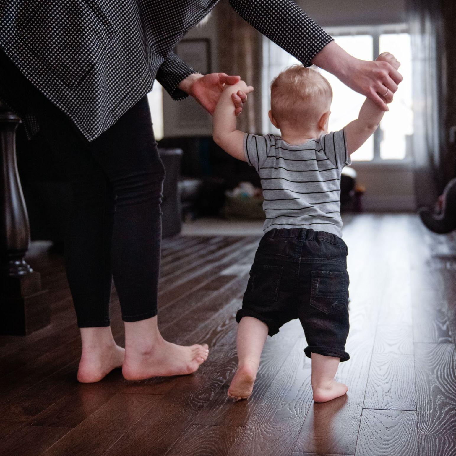Ein Kleinkind lernt in einem Wohnzimmer an den Händen seiner Mutter laufen.