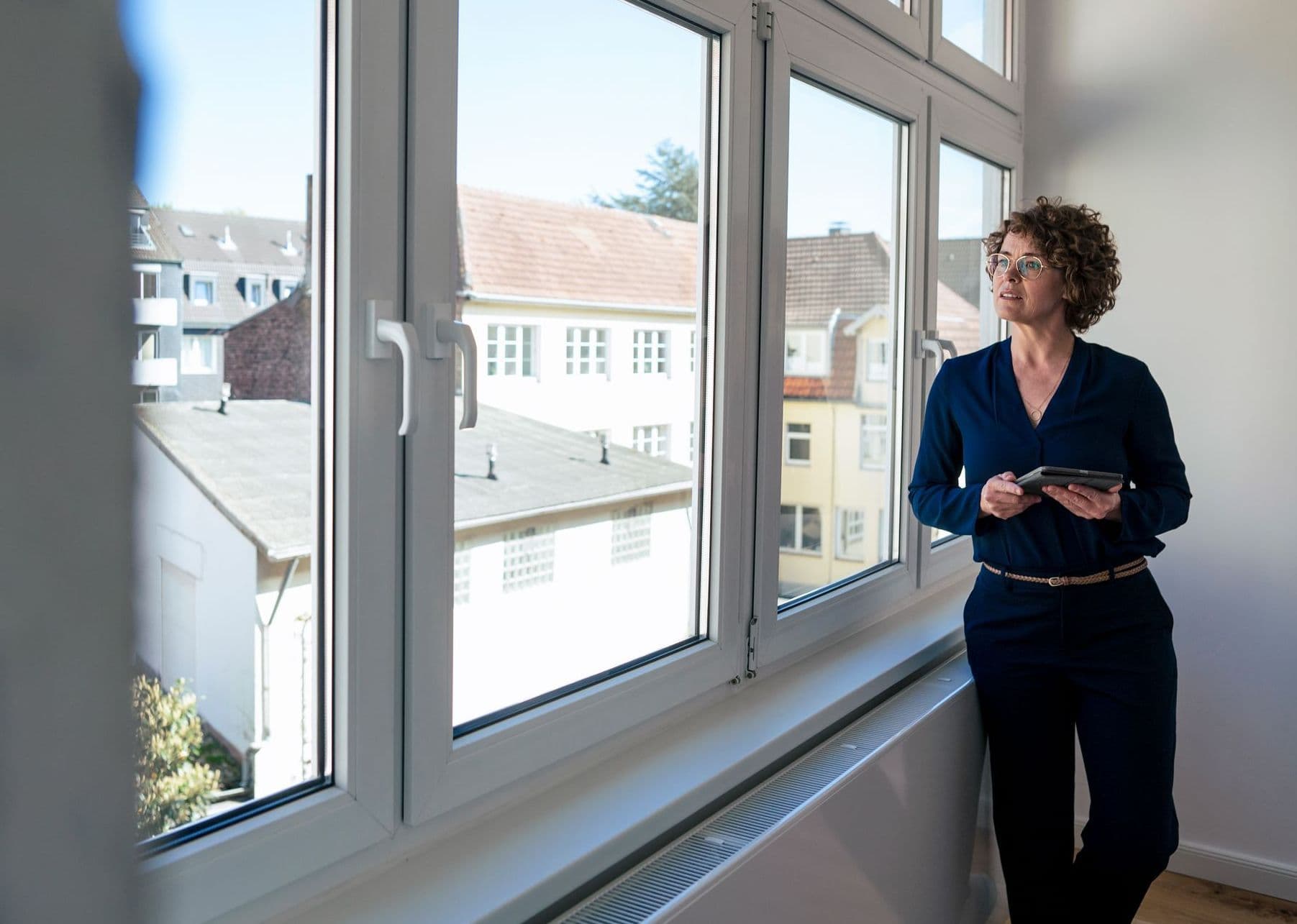 Eine Frau steht in der Ecke eines Zimmers angelehnt an ein Fenster und schaut hinaus.