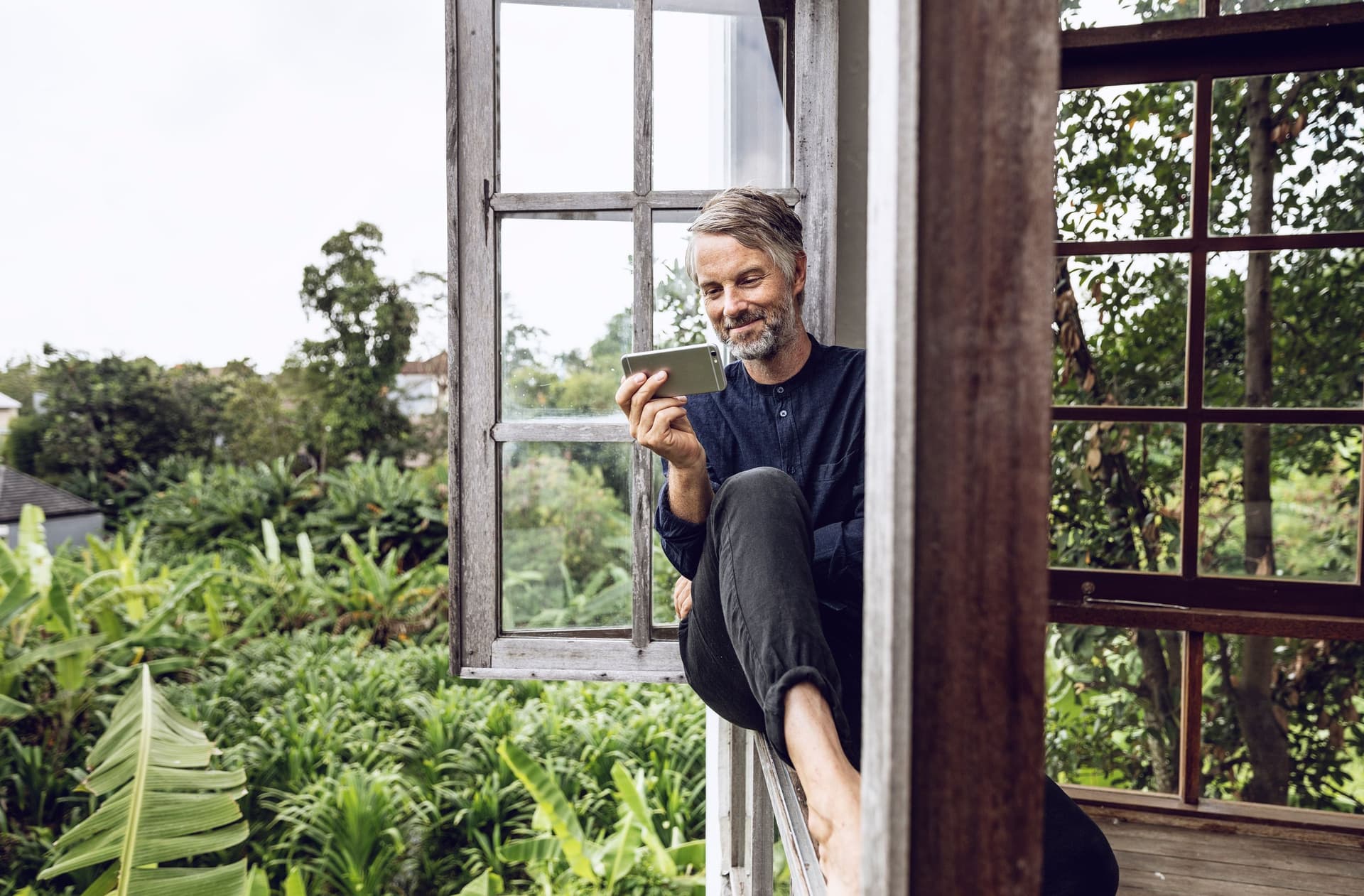 Ein Mann mittleren Alters sitzt in einem Fenster über einem Garten und schaut lächelnd auf sein Smartphone. 