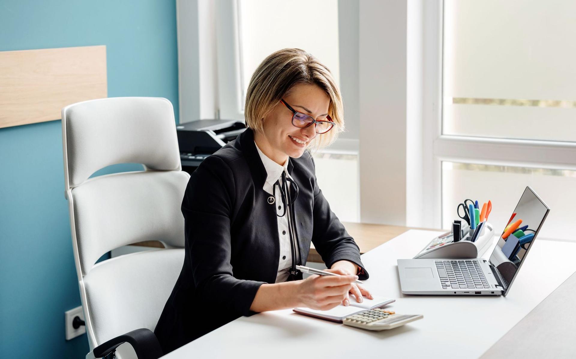 Eine lächelnde Geschäftsfrau mit Brille schreibt in ein Notizbuch an einem hellen Bürotisch mit Laptop und Büromaterialien.