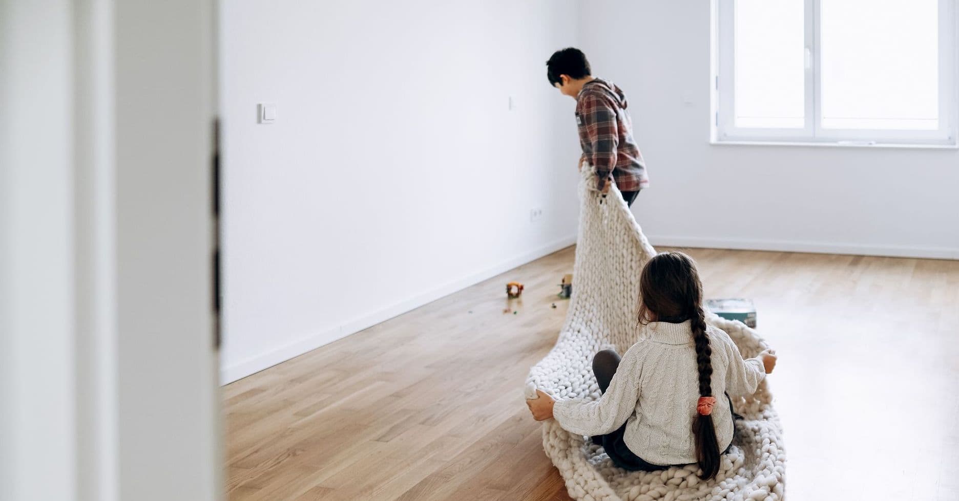 Zwei Kinder spielen in einem leeren Zimmer, eines sitzt auf einer großen Decke, das andere zieht sie über den Holzfußboden