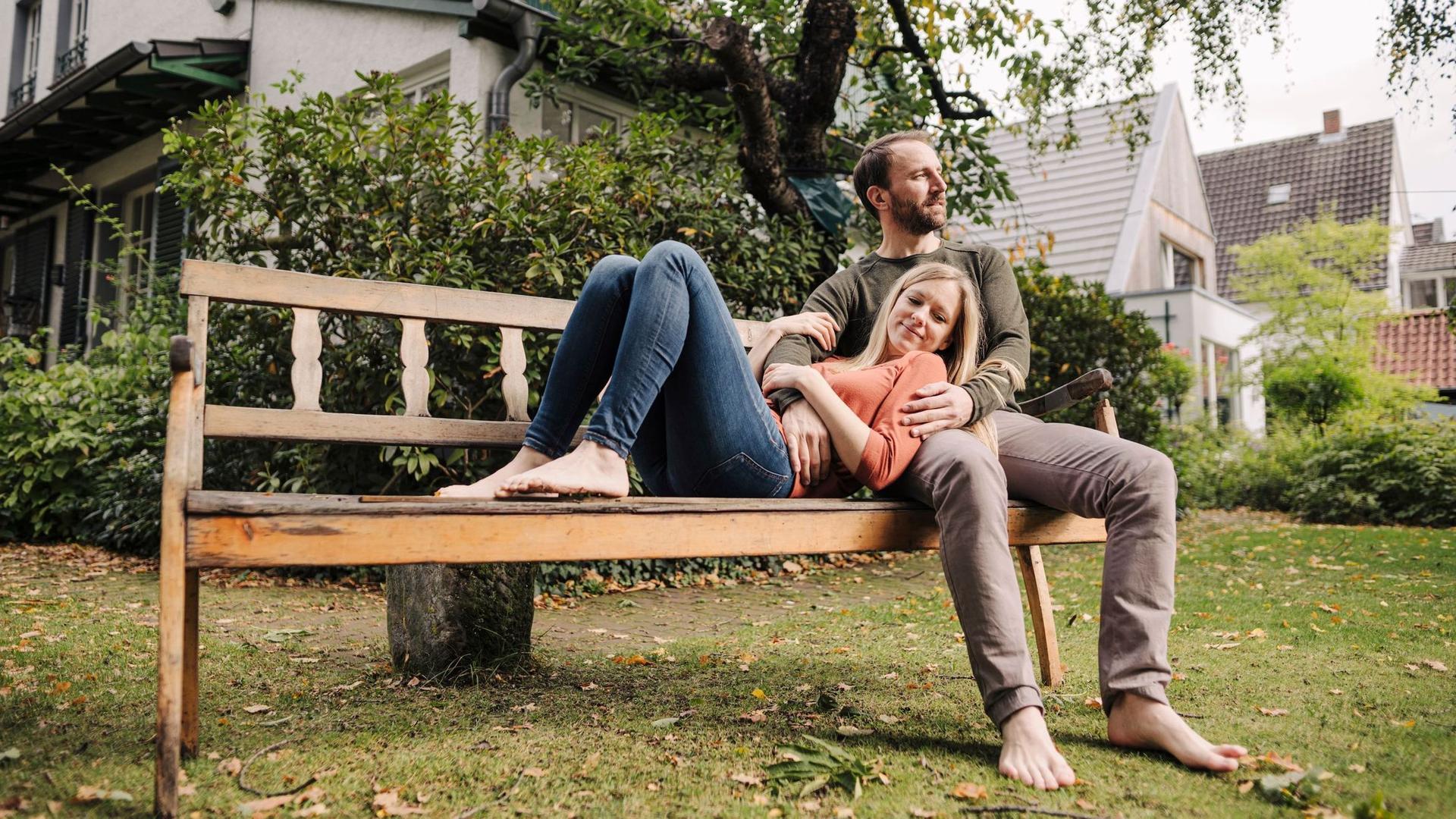 Ein Paar entspannt auf einer Parkbank im Grünen vor Häusern, die Frau lehnt sich an den Mann.