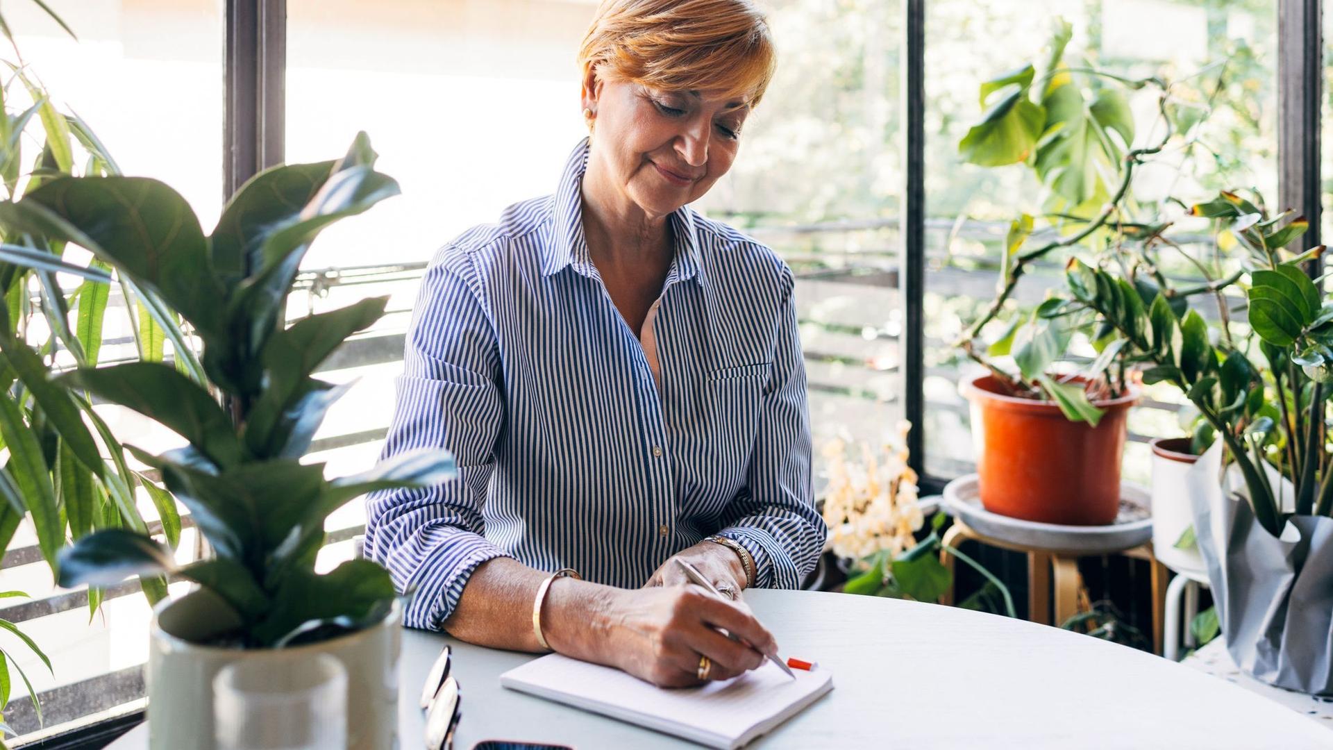 Eine ältere Frau schreibt lächelnd in ein Notizbuch an einem pflanzenreichen, sonnigen Tisch.