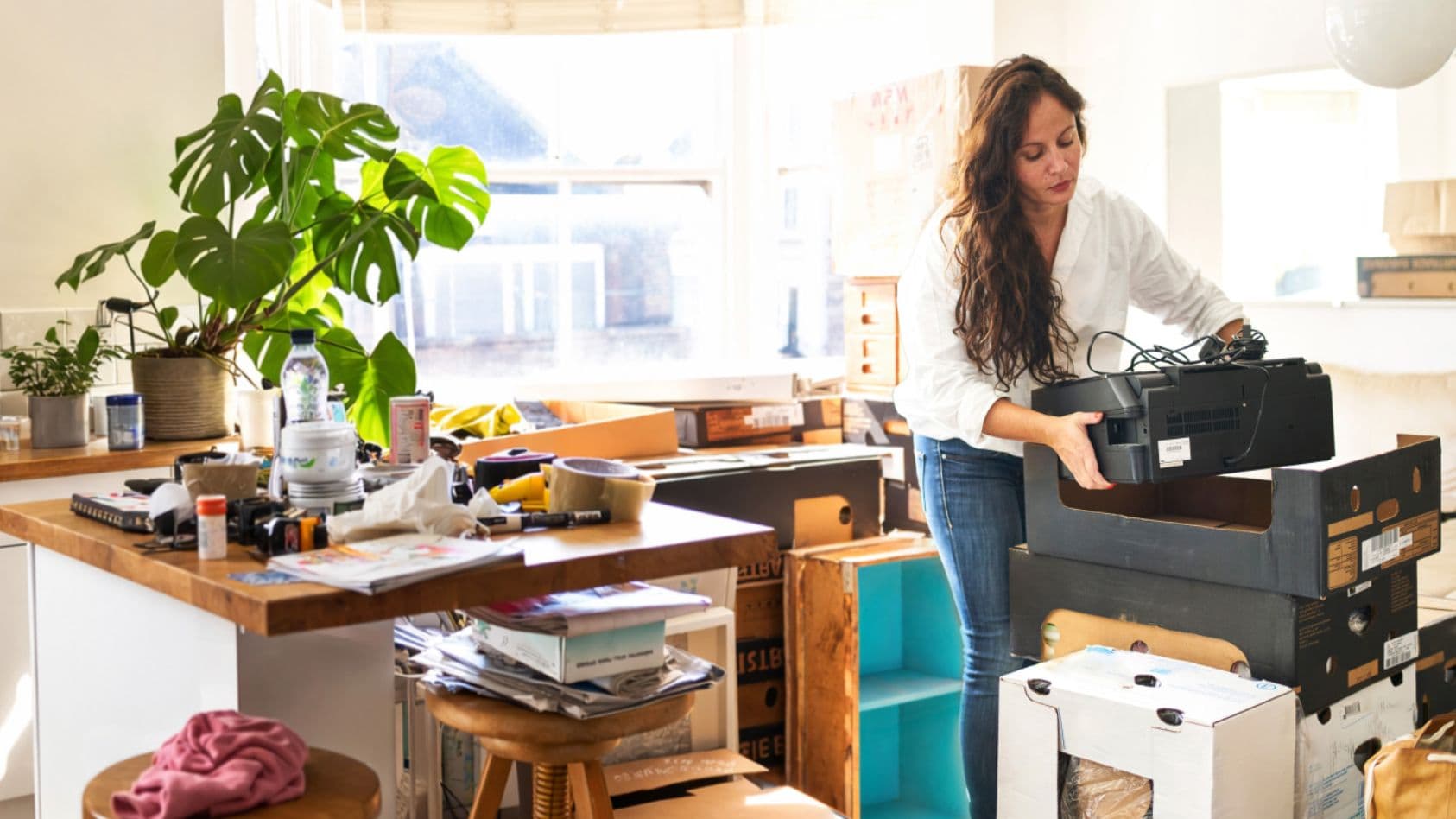 Frau packt Umzugskisten in helle, unordentliche Küche während Sonnenlicht durchs Fenster fällt