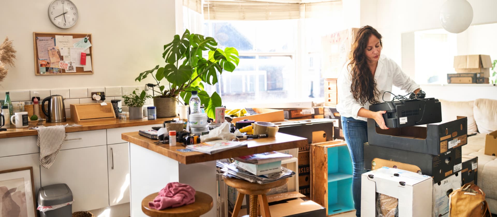 Frau packt Umzugskisten in helle, unordentliche Küche während Sonnenlicht durchs Fenster fällt