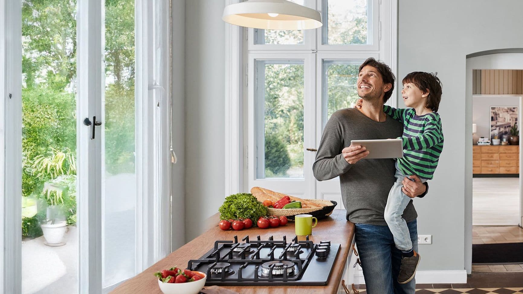 Vater hält Kind im Arm in moderner Küche mit Tablet in der Hand