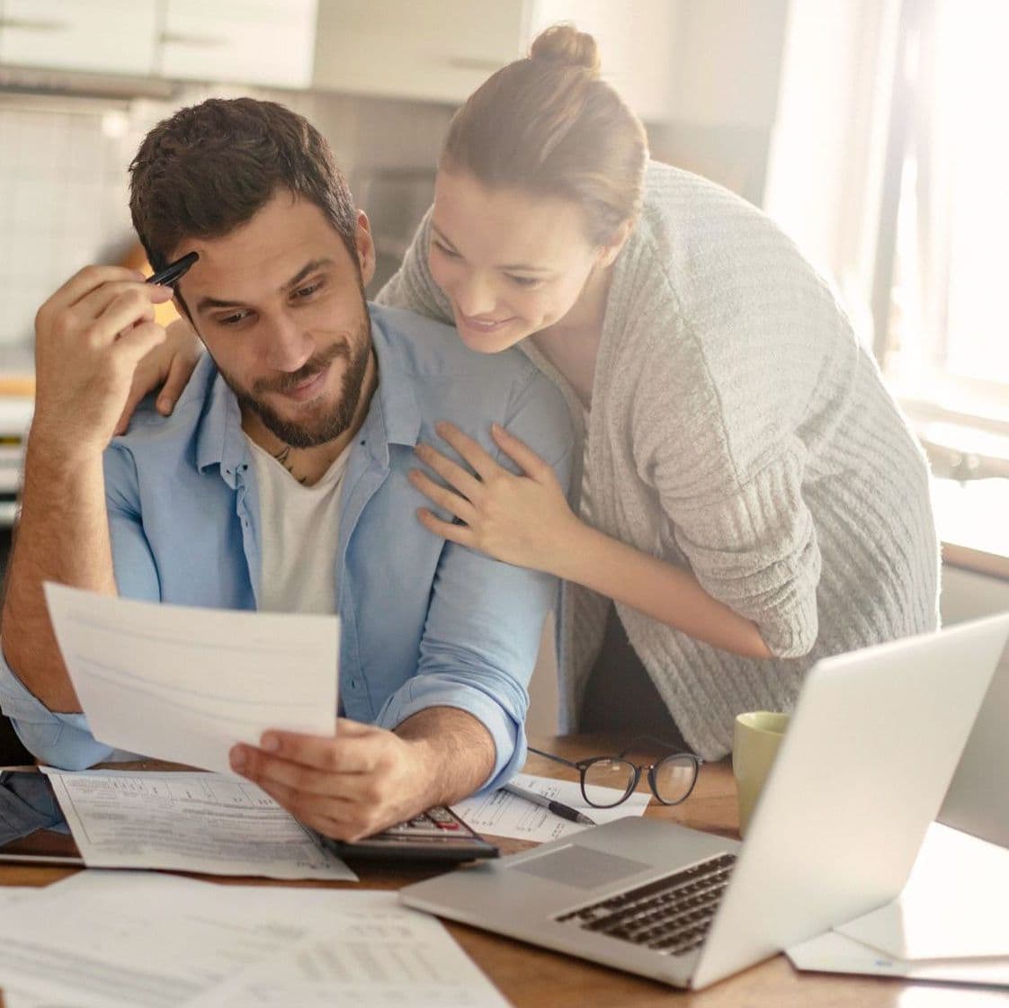 junges paar sitzt in heller küche und prüft gemeinsam unterlagen auf einem tisch mit laptop und kaffeekanne