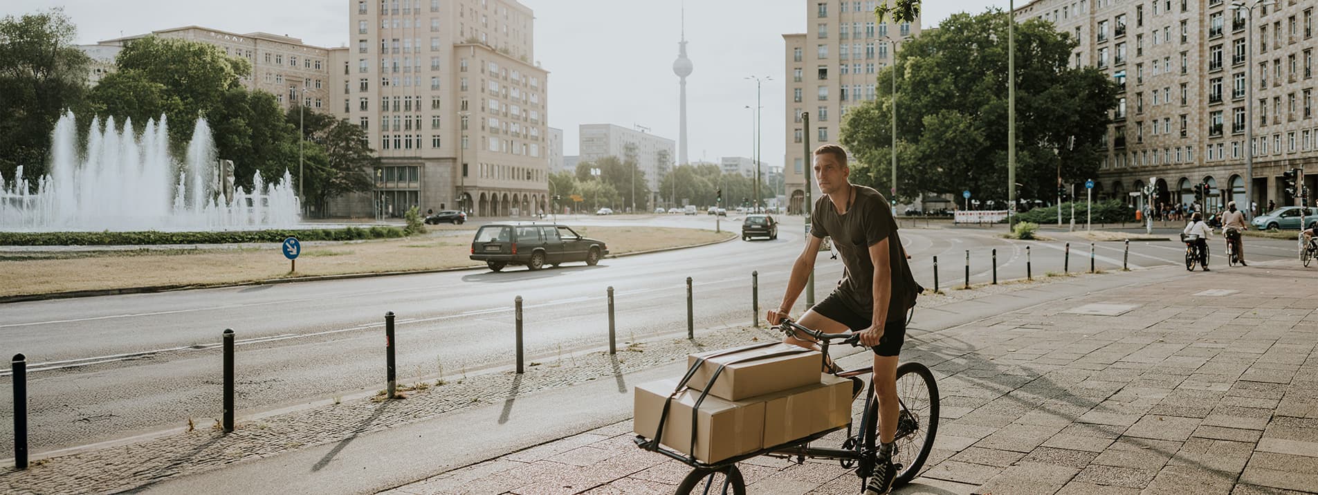 Mann auf Lastenfahrrad mit Paketen fährt an einer Stadtstraße vorbei, im Hintergrund Springbrunnen und Fernsehturm von Berlin.