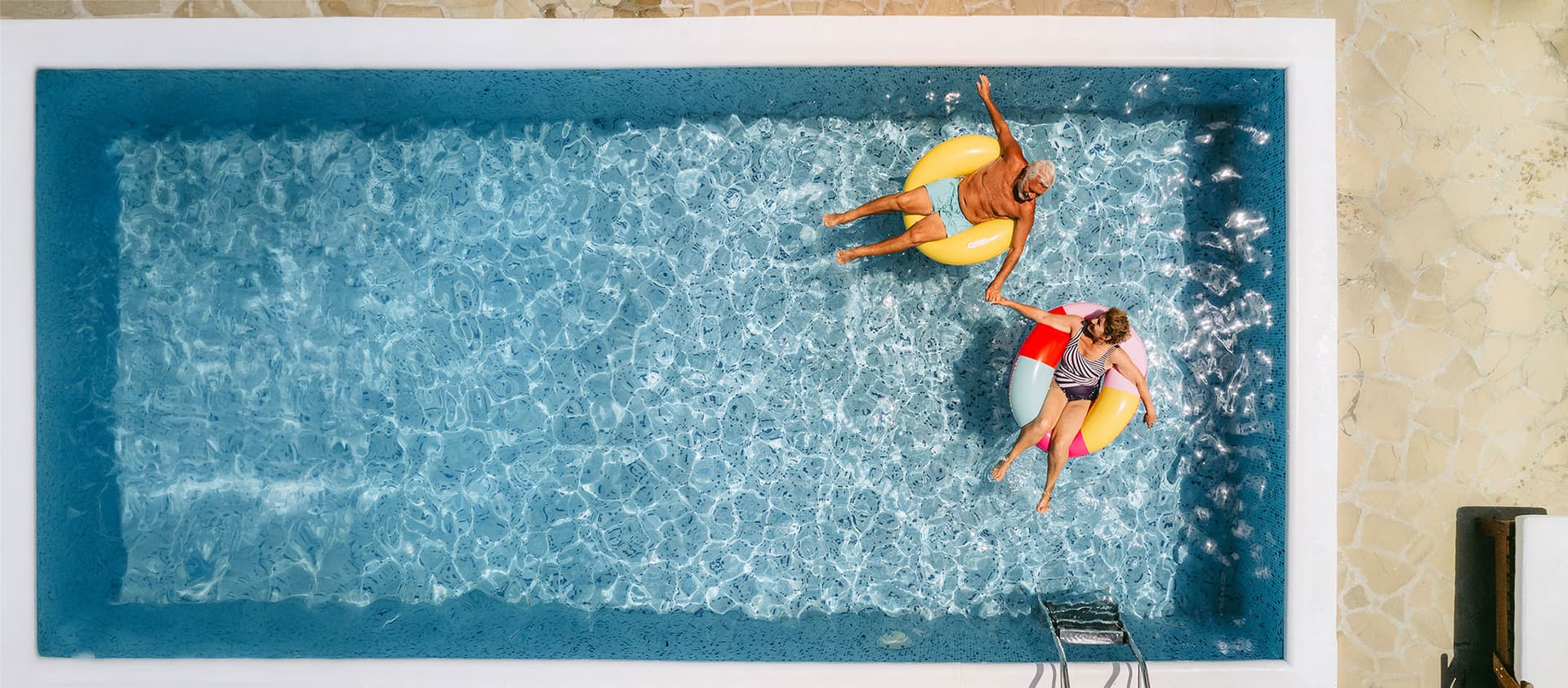 Luftaufnahme von zwei älteren Personen, die auf bunten Schwimmringen in einem rechteckigen Pool treiben und sich an den Händen halten.