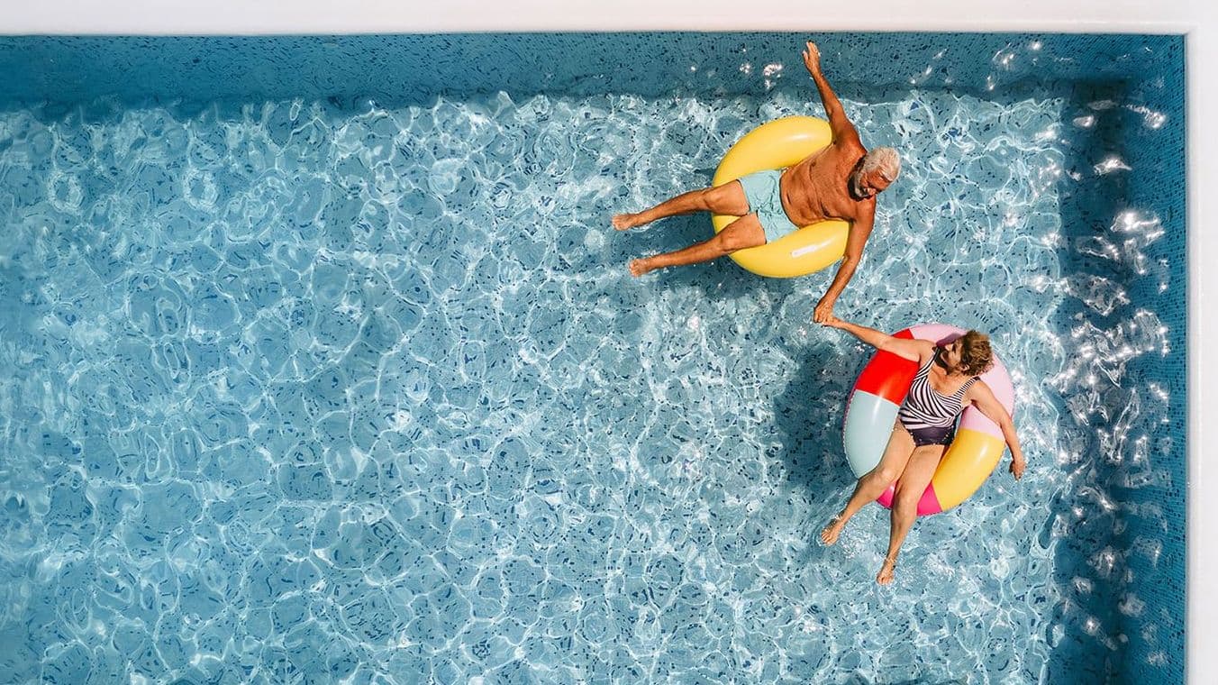 Luftaufnahme von zwei älteren Personen, die auf bunten Schwimmringen in einem rechteckigen Pool treiben und sich an den Händen halten.