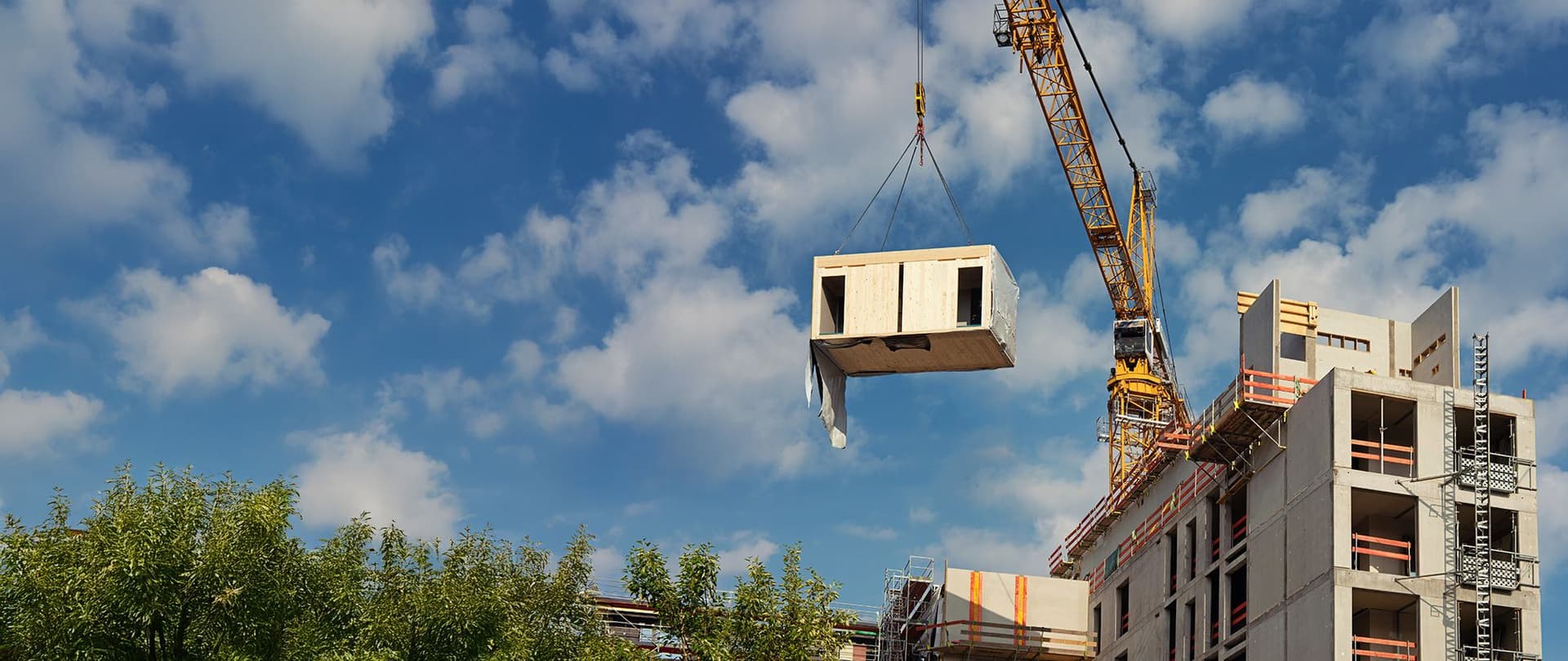 Baukran hebt ein vorgefertigtes Betonmodul an und setzt es auf ein mehrstöckiges Gebäude im Rohbau unter blauem Himmel