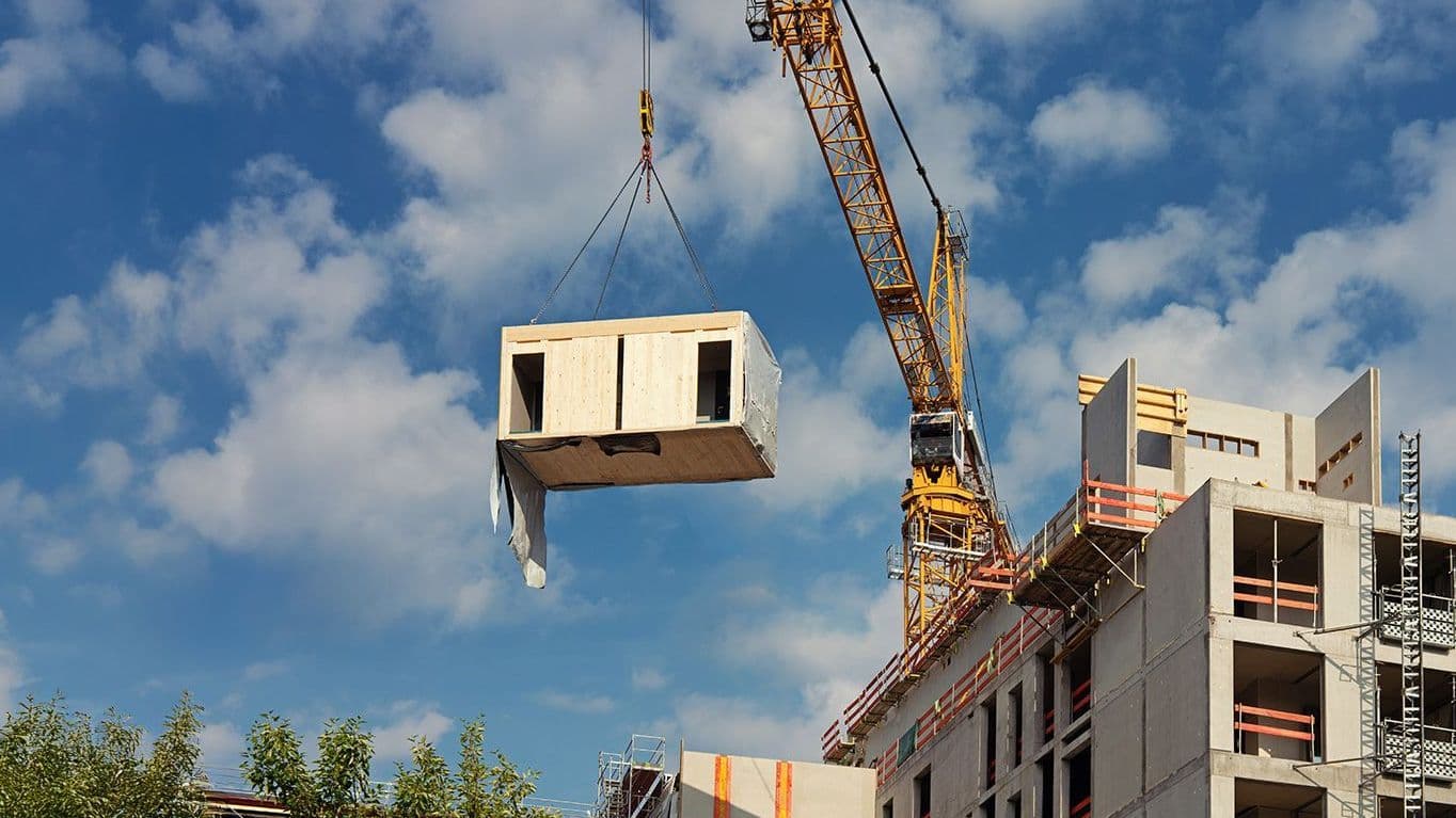 Baukran hebt ein vorgefertigtes Betonmodul an und setzt es auf ein mehrstöckiges Gebäude im Rohbau unter blauem Himmel