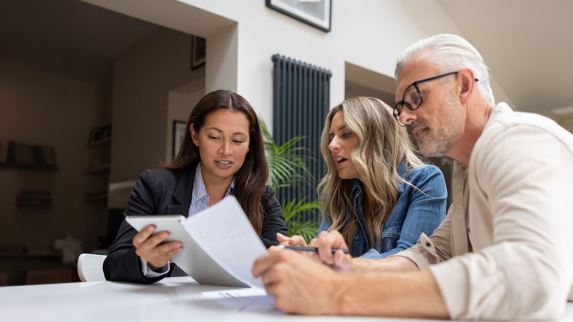 Drei Personen sitzen an einem Tisch und besprechen Dokumente, eine Person hält ein Tablet, moderne Innenraumatmosphäre mit natürlichem Licht