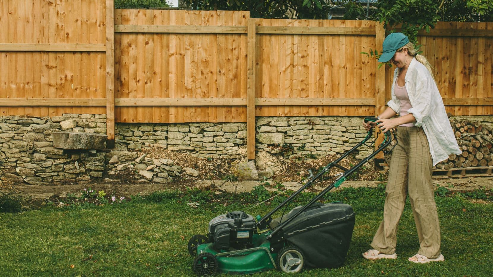 Person mit grüner Baseballkappe mäht Rasen im Garten mit Holzzaun und Steinmauer im Hintergrund.