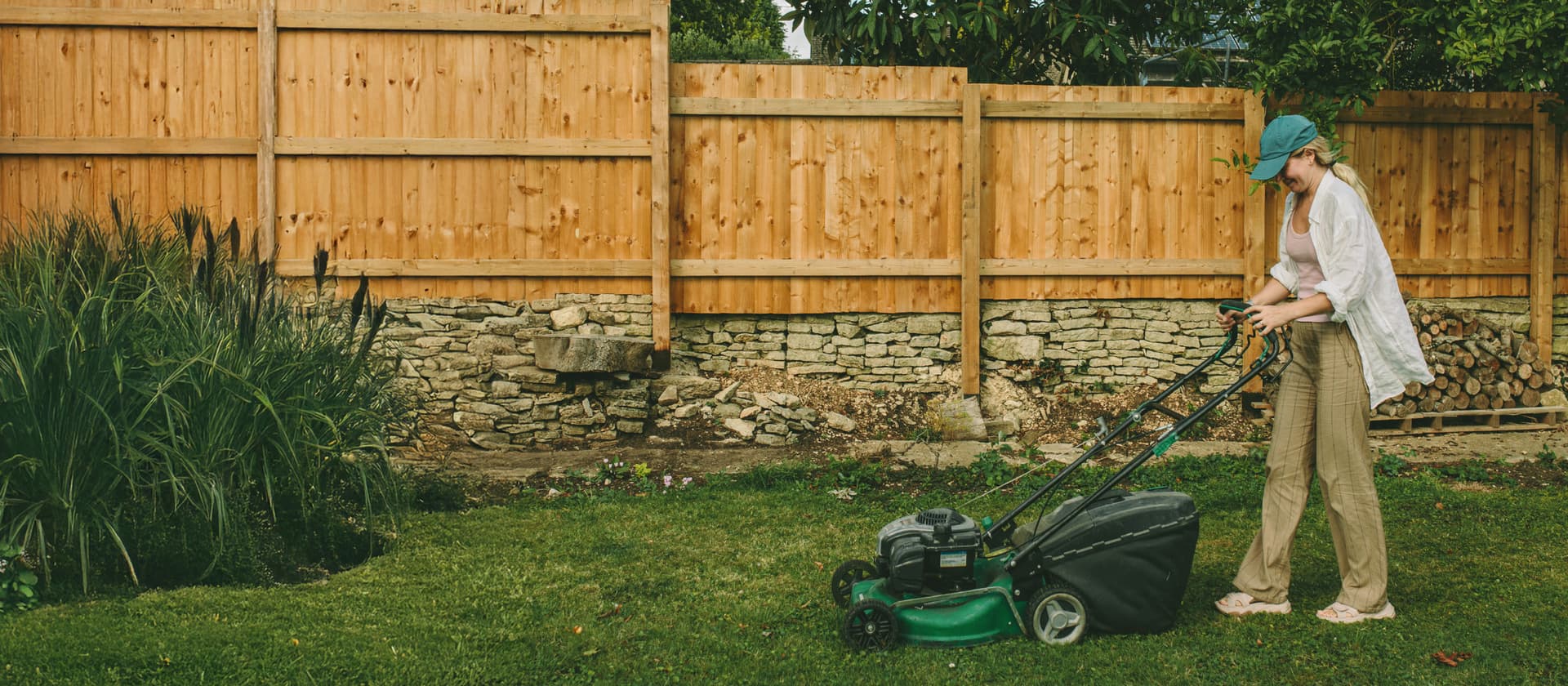 Person mit grüner Baseballkappe mäht Rasen im Garten mit Holzzaun und Steinmauer im Hintergrund.