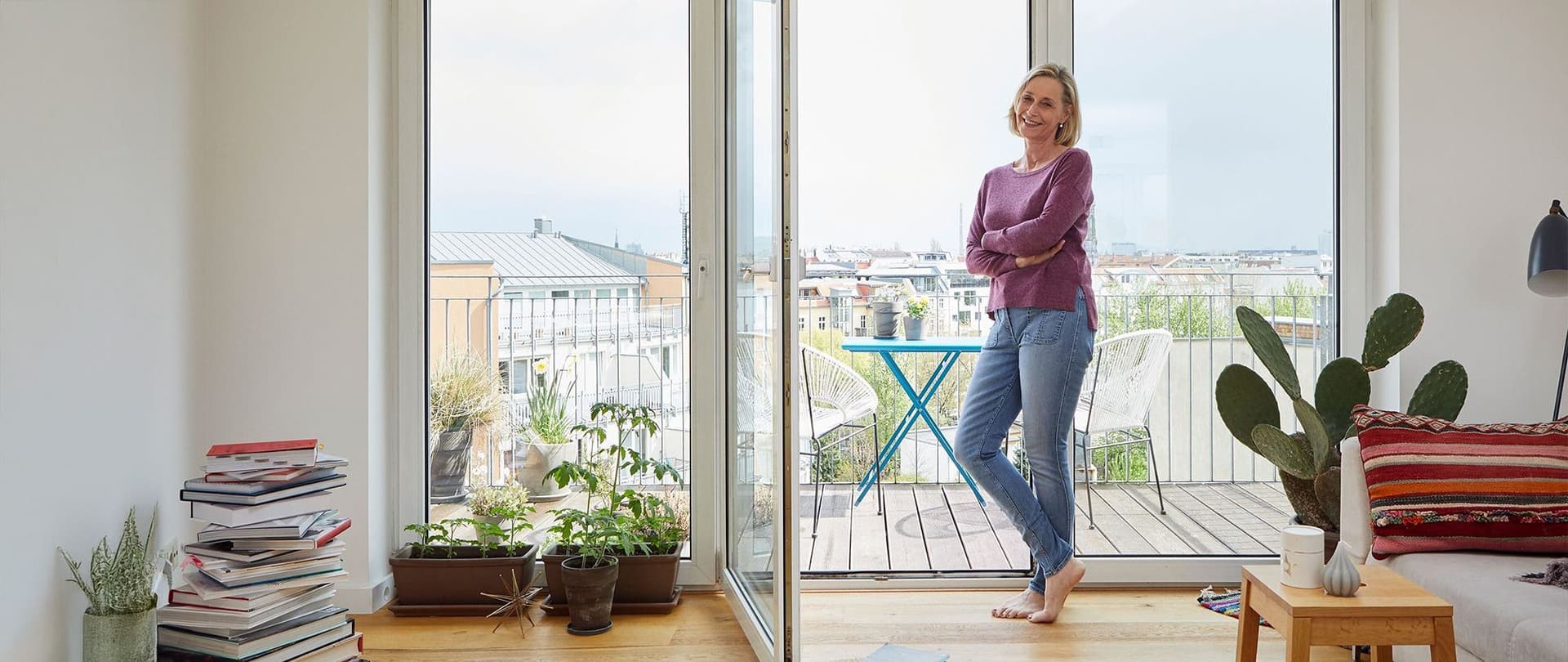 Frau steht barfuß in einem hellen Wohnzimmer vor geöffneter Balkontür mit Blick auf Stadt und Terrasse