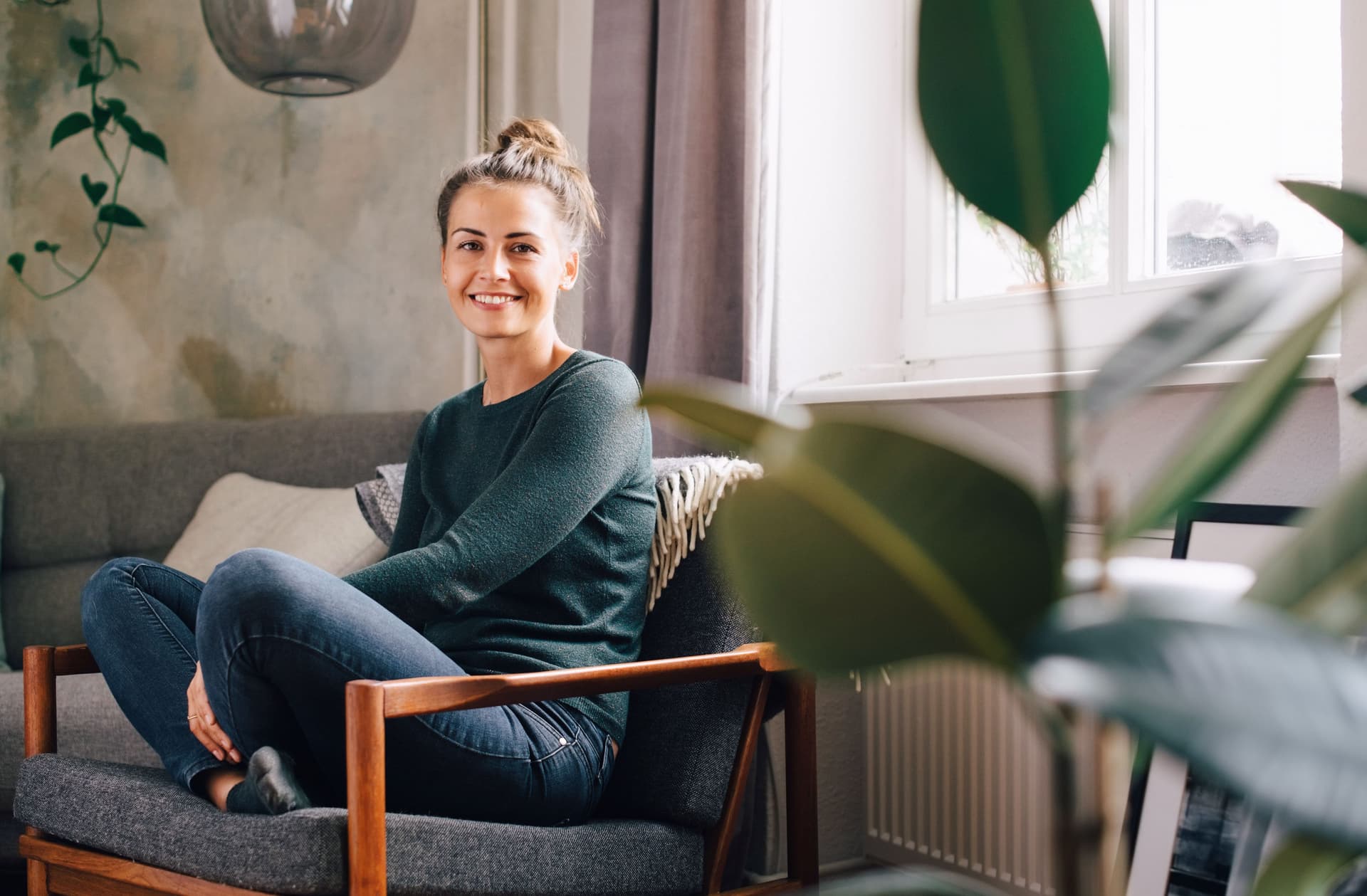 Eine blonde Frau in grünem Pulli sitzt lächelnd im Schneidersitz auf einem Sofa.