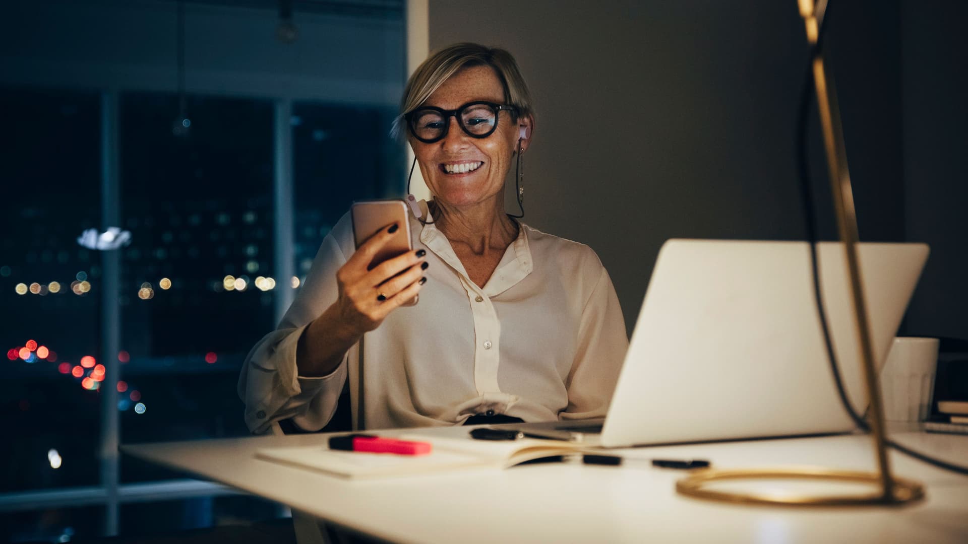 Lächelnde Frau sitzt mit Smartphone in der Hand an einem Schreibtisch. Ihr Laptop steht auf dem Tisch. Durch das Fenster sieht man Lichter einer Stadt, es ist dunkel.