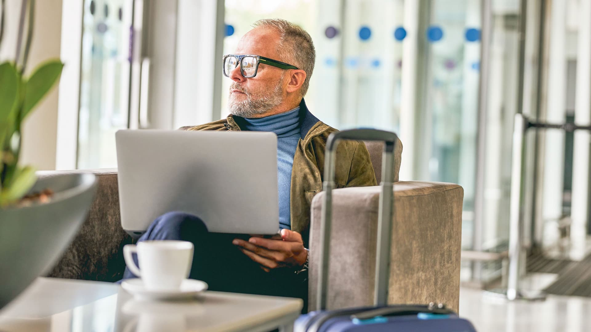 Älterer Herr sitzt mit Laptop auf dem Schoß vor Glasfront in einem Lounge-Bereich und schaut raus.