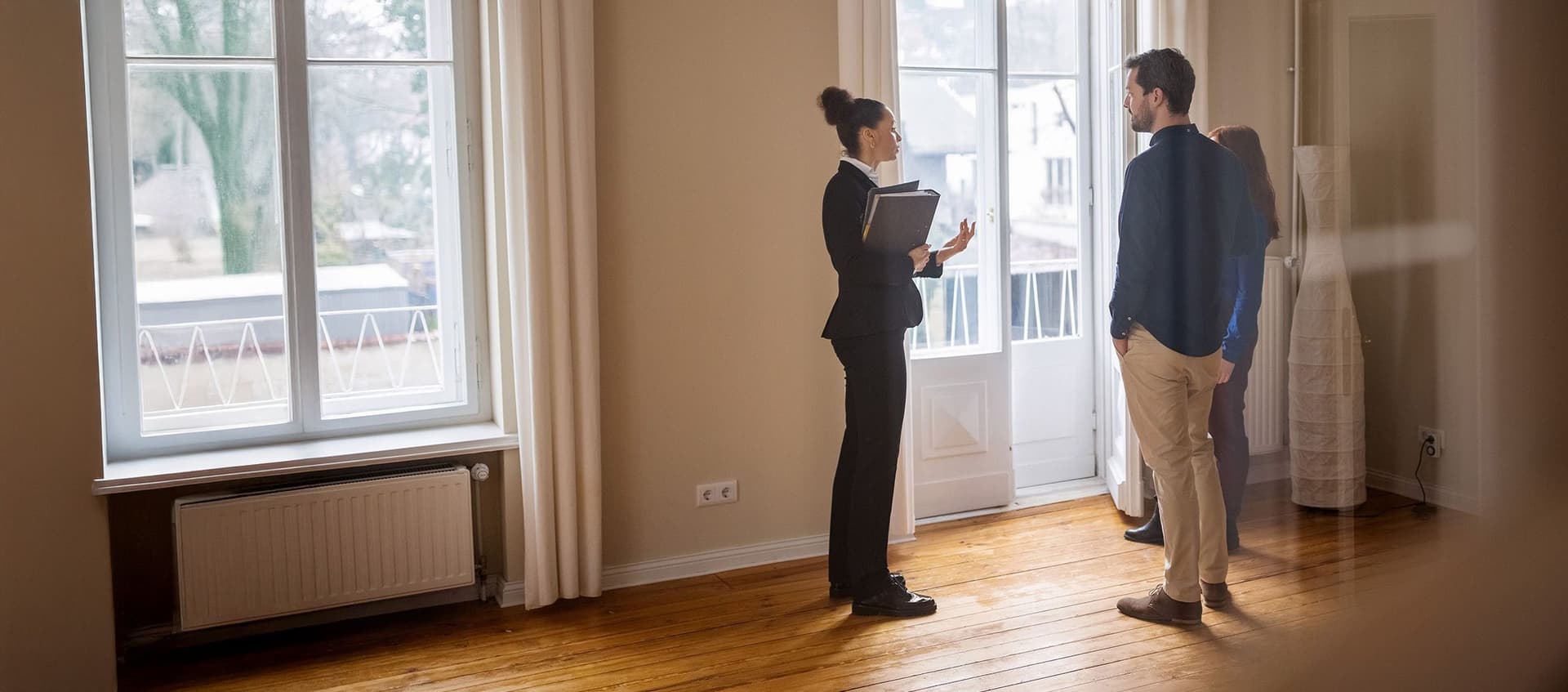 Immobilienmaklerin steht mit einem jungen Paar in einem leeren Zimmer. Sie trägt einen Hosenanzug und hat eine Mappe in der Hand.