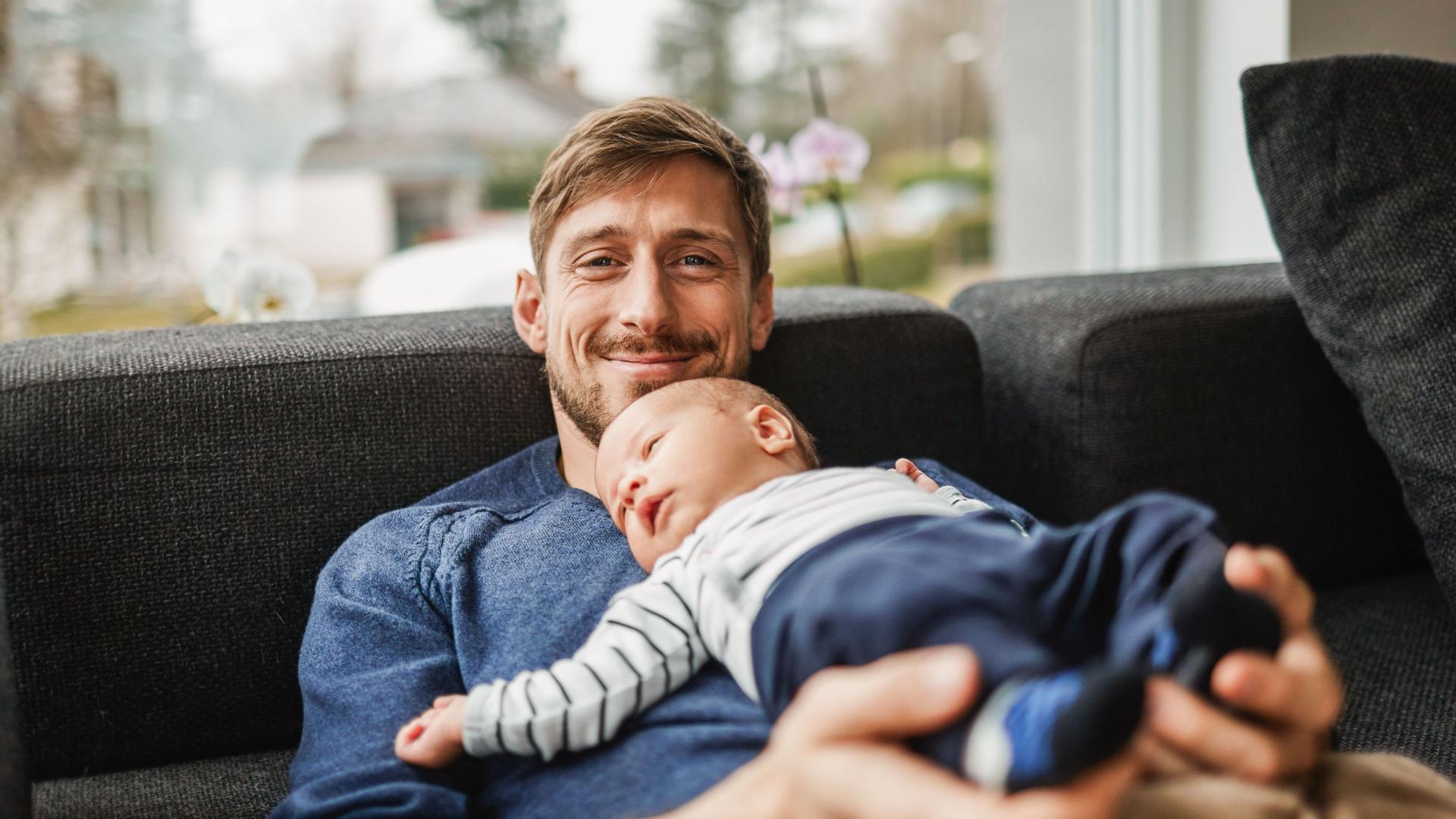 Ein Mann sitzt mit seinem Baby glücklich in seinem Haus auf der Couch.
