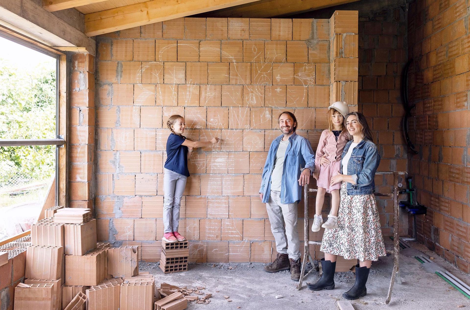 Eine vierköpfige Familie steht in dem Rohbau ihres neuen Hauses vor einer unverputzten Mauersteinwand und schaut in die Kamera. 