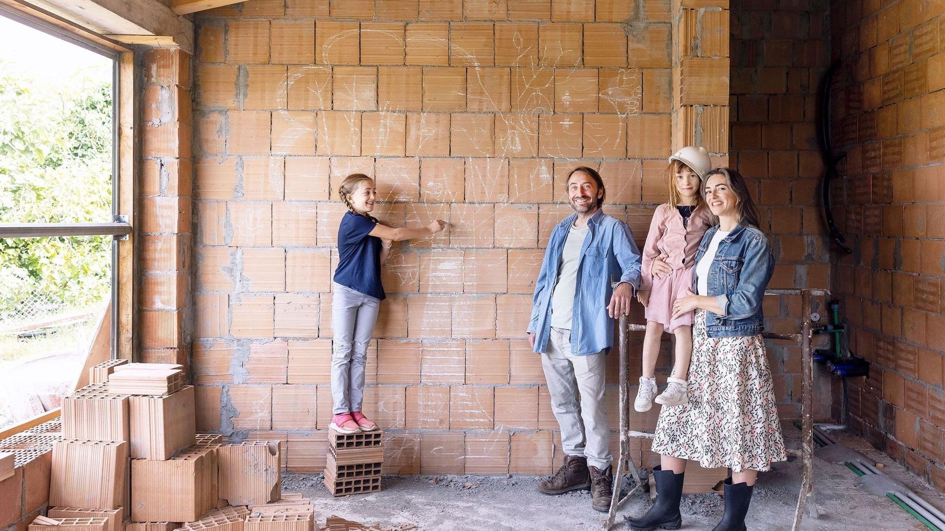 Eine vierköpfige Familie steht in dem Rohbau ihres neuen Hauses vor einer unverputzten Mauersteinwand und schaut in die Kamera. 