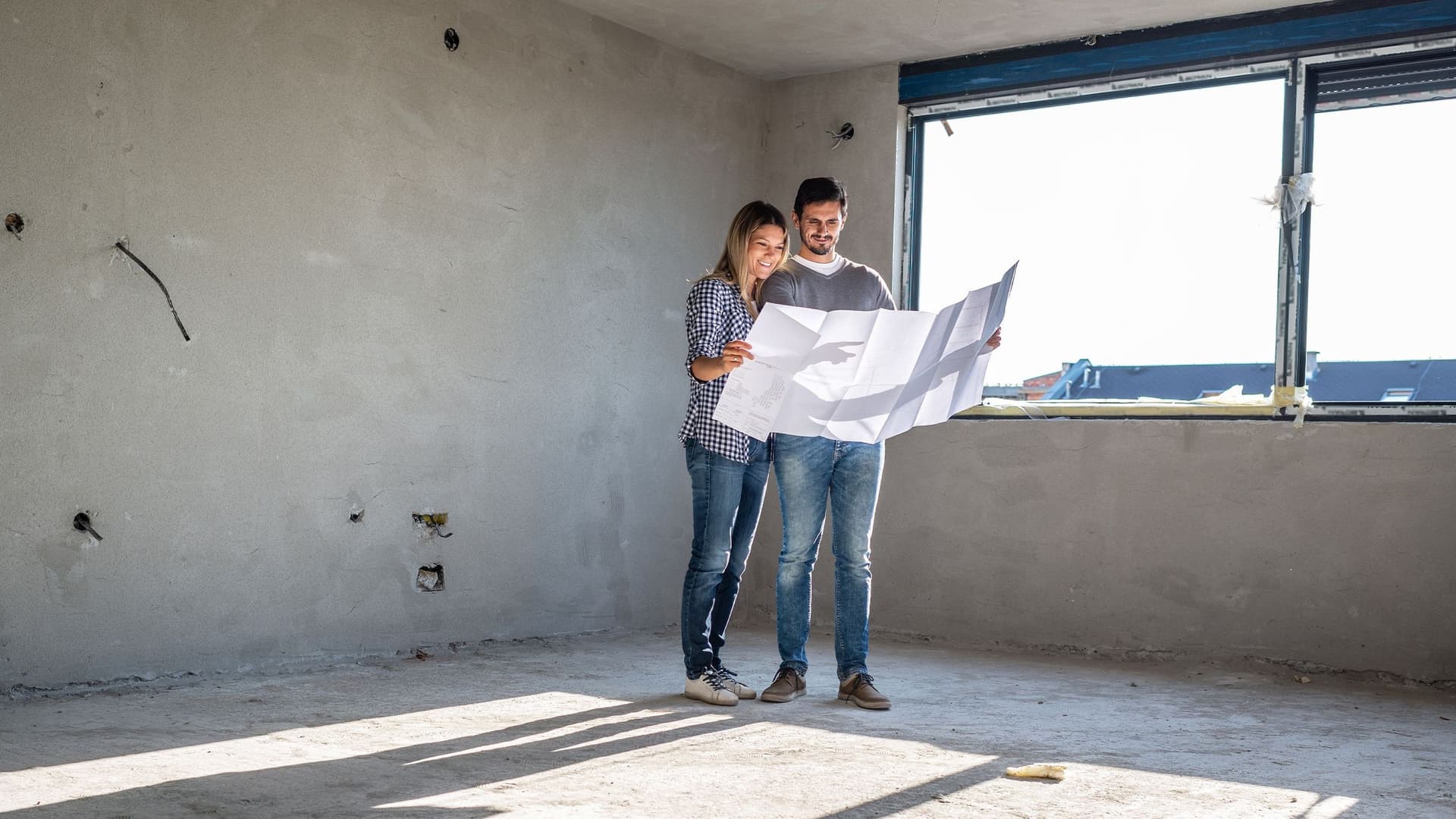 Eine Frau und ein Mann stehen inmitten eines Rohbaus auf einer Baustelle und sehen sich zusammen den Bauplan an. 