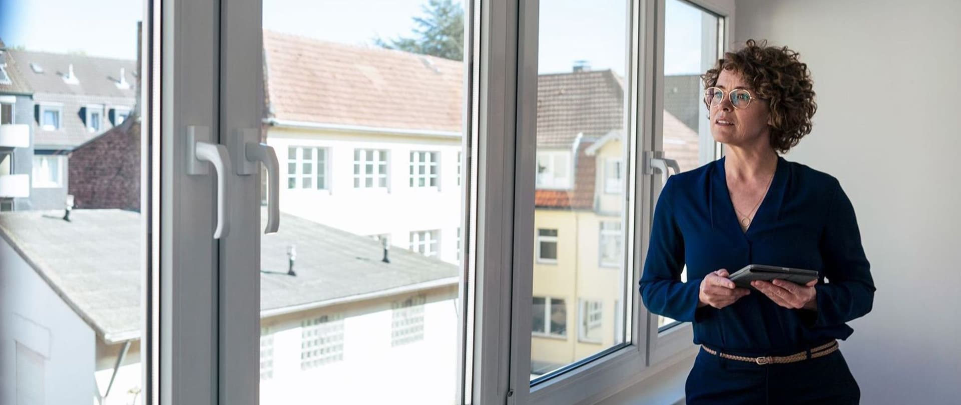 Eine Frau steht in der Ecke eines Zimmers angelehnt an ein Fenster und schaut hinaus.