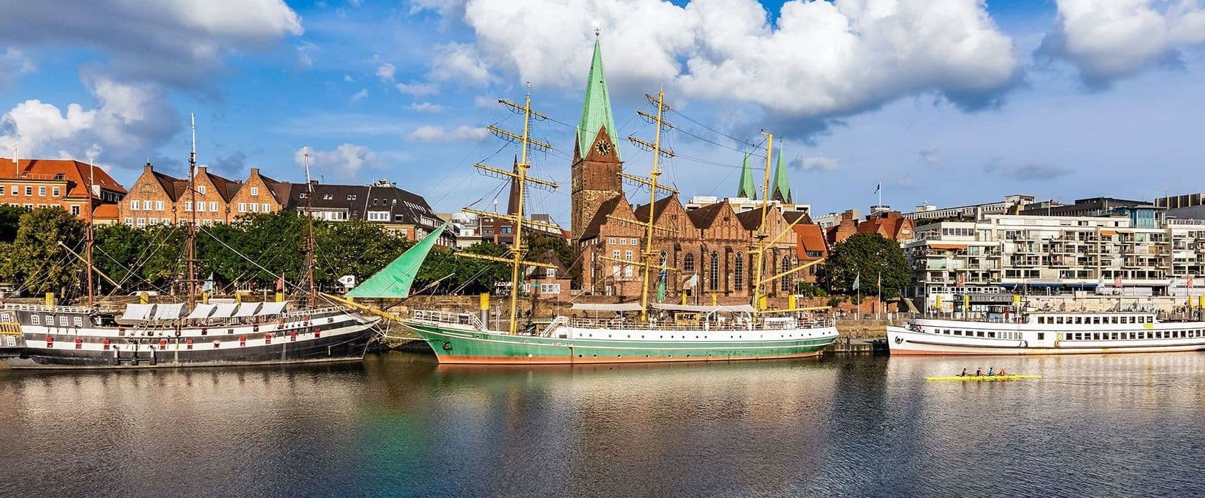 Hafenansicht mit mehreren angelegten Segelschiffen und einem Ruderboot im Vordergrund vor historischen Backsteinbauten und einer Kirche mit grünem Turm.