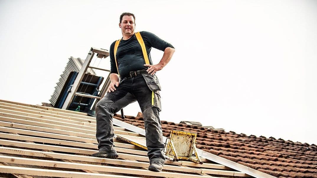 Ein Dachdecker steht selbstbewusst auf einem teilweise gedeckten Dach. Er trägt Arbeitskleidung mit einem schwarzen Shirt und gelben Hosenträgern. Im Hintergrund ist eine Leiter und weiteres Werkzeug zu sehen, das er für seine Arbeit benötigt.