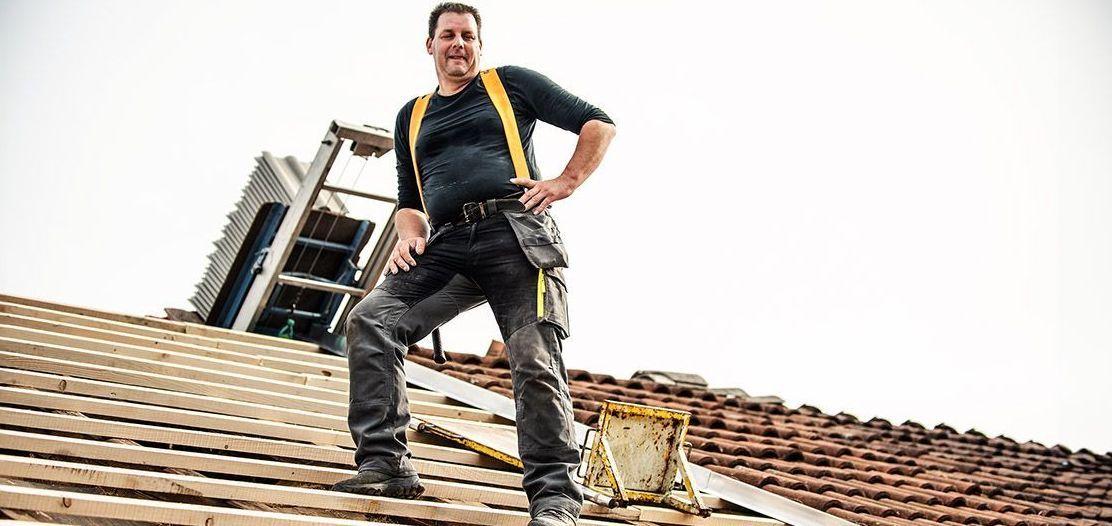Ein Dachdecker steht selbstbewusst auf einem teilweise gedeckten Dach. Er trägt Arbeitskleidung mit einem schwarzen Shirt und gelben Hosenträgern. Im Hintergrund ist eine Leiter und weiteres Werkzeug zu sehen, das er für seine Arbeit benötigt.