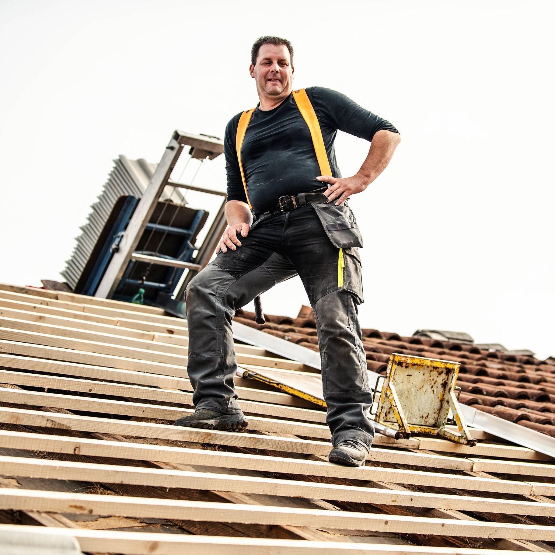 Ein Dachdecker steht selbstbewusst auf einem teilweise gedeckten Dach. Er trägt Arbeitskleidung mit einem schwarzen Shirt und gelben Hosenträgern. Im Hintergrund ist eine Leiter und weiteres Werkzeug zu sehen, das er für seine Arbeit benötigt.