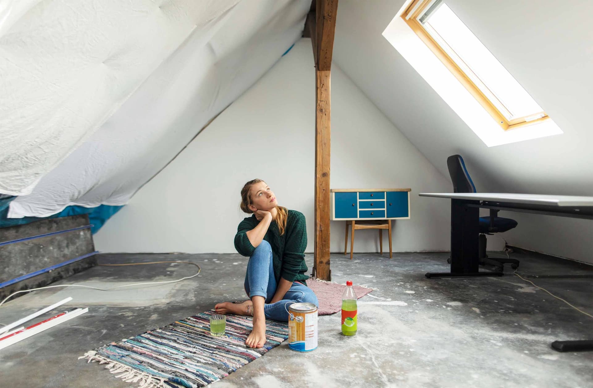 Frau sitzt barfuß in einem unausgebauten Dachgeschoss auf Teppich mit Getränk und Farbeimer, nachdenklicher Blick Richtung Dachfenster