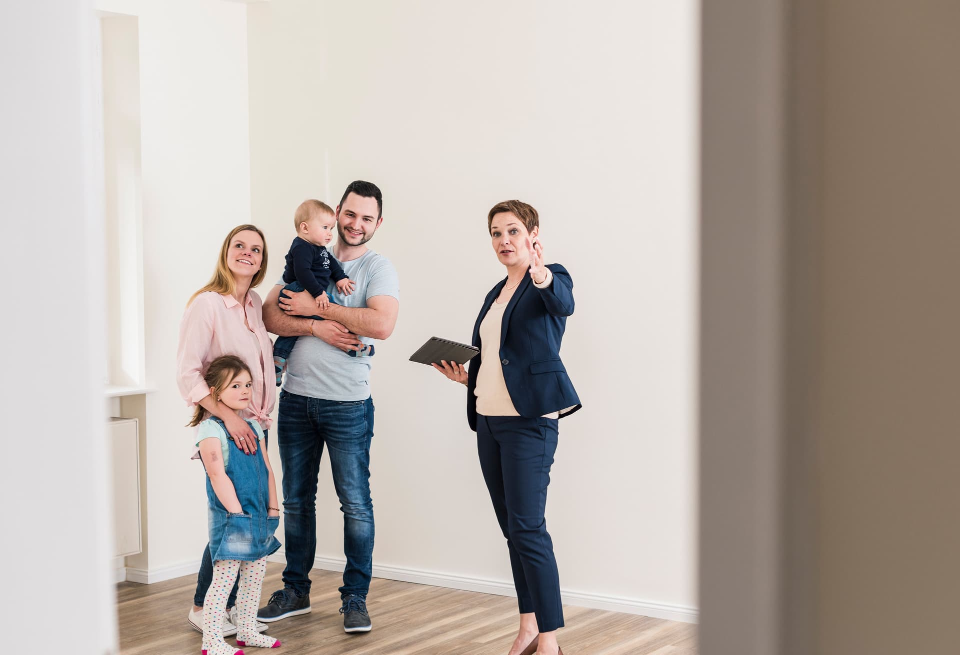 Eine Familie mit zwei Kindern wird von einer Maklerin in einer leeren Wohnung herumgeführt.