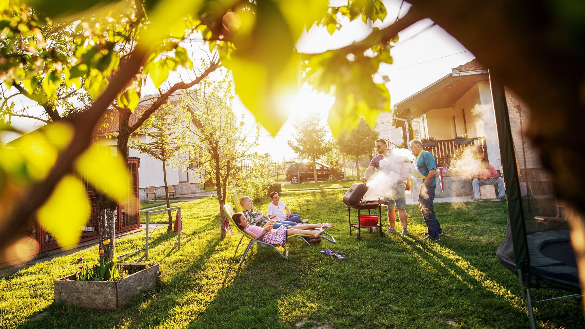 Familie grillt im Garten umgeben von kleineren Bäumen. Die Sonne scheint in die Kamera.