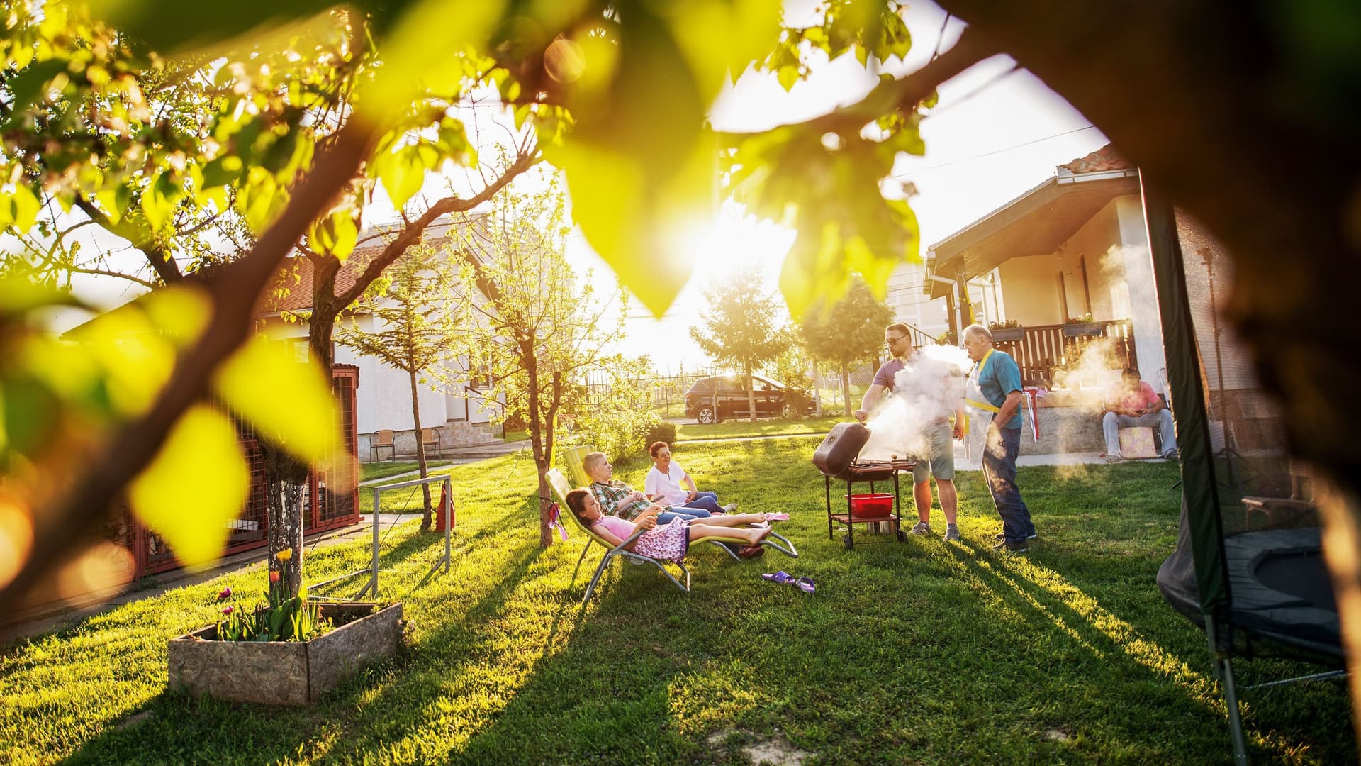 Familie grillt im Garten umgeben von kleineren Bäumen. Die Sonne scheint in die Kamera.