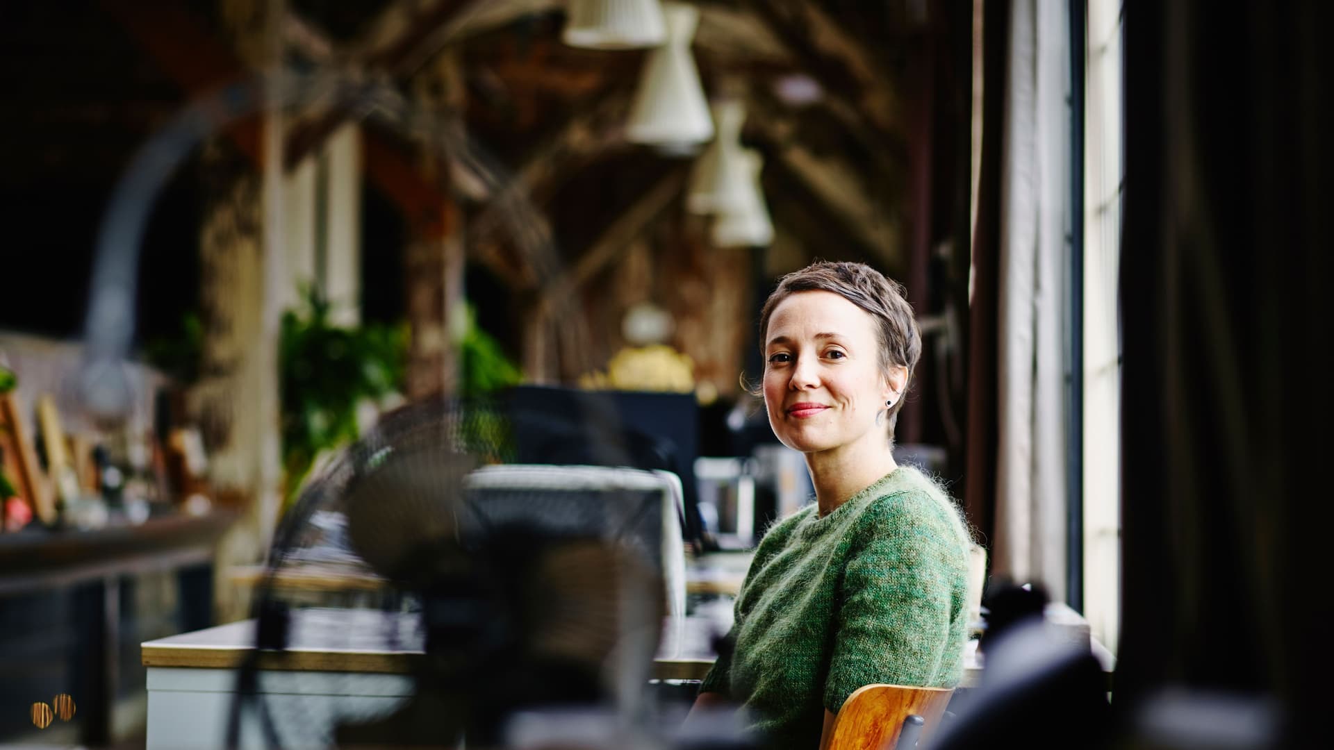 Frau, die an einem Tisch in einem Design-Büro sitzt und in die Kamera lächelt.