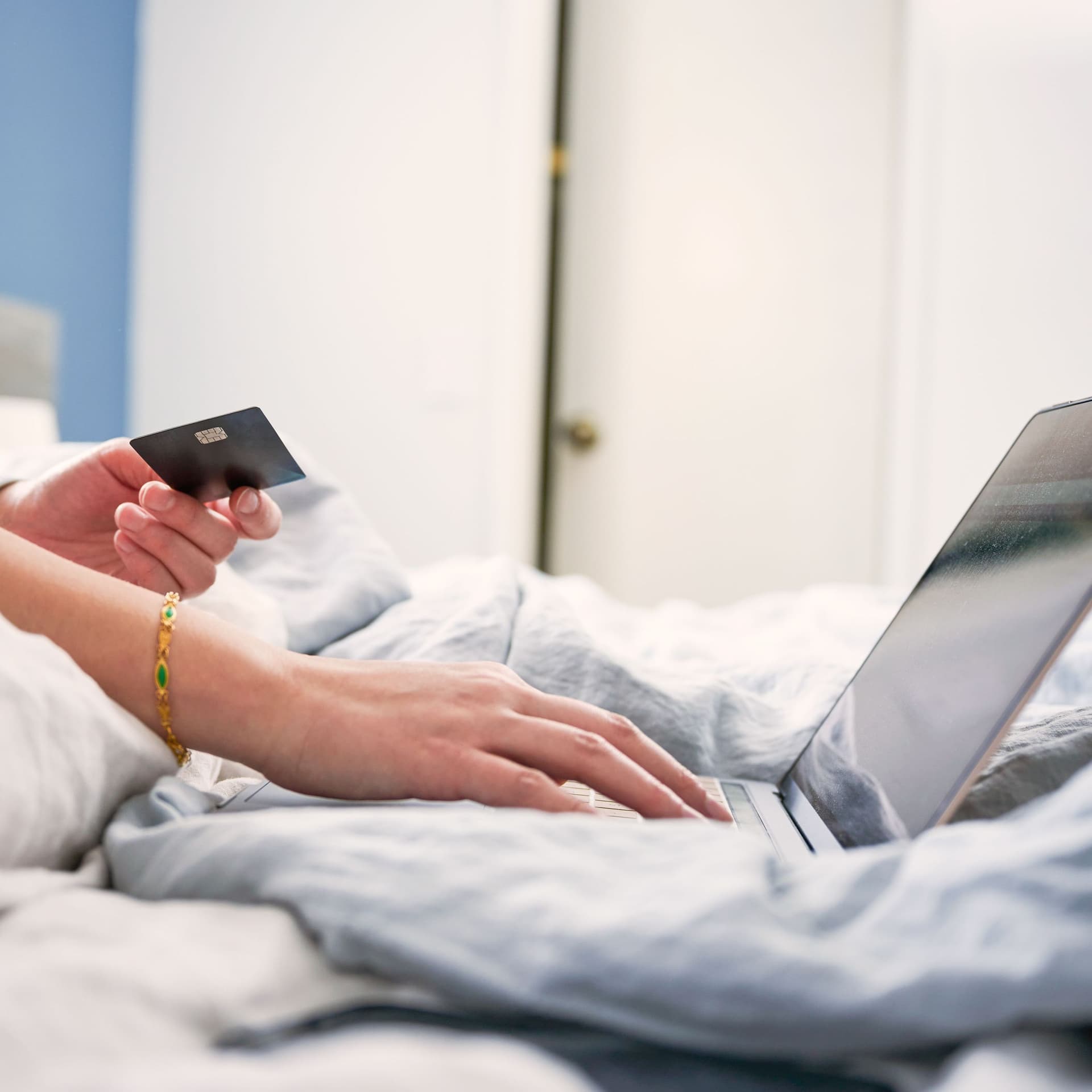 Nahaufnahme einer Frau im Bett sitzend mit einer Bankkarte in der Hand. Mit der anderen bedient sie einen Laptop.