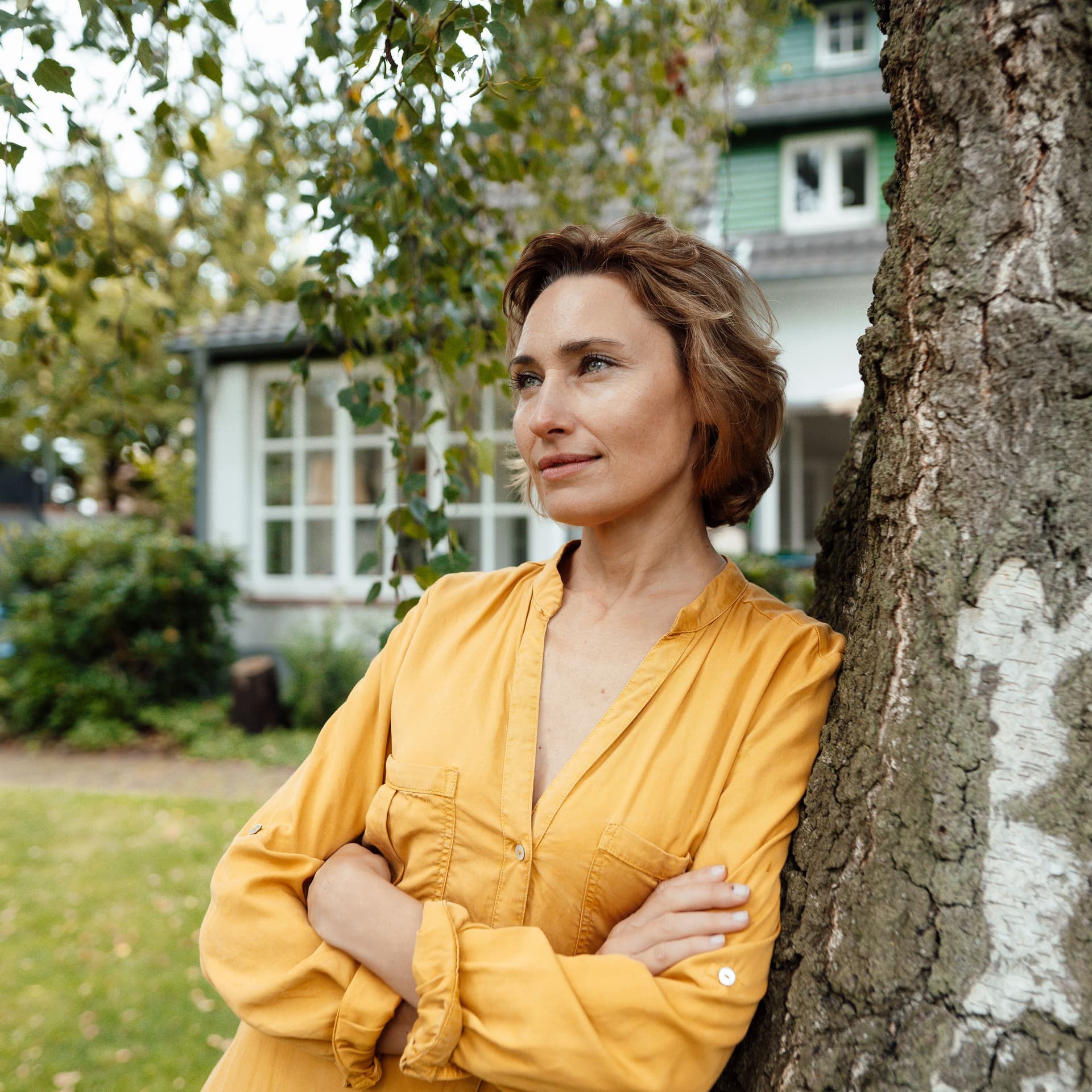 frau-lehnt-an-baum-vor-ihrem-haus-GI-1404450086
