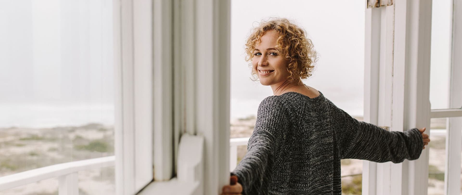 Frau mit lockigen Haaren öffnet die Balkontür eines Strandhauses und lächelt zurück in die Kamera