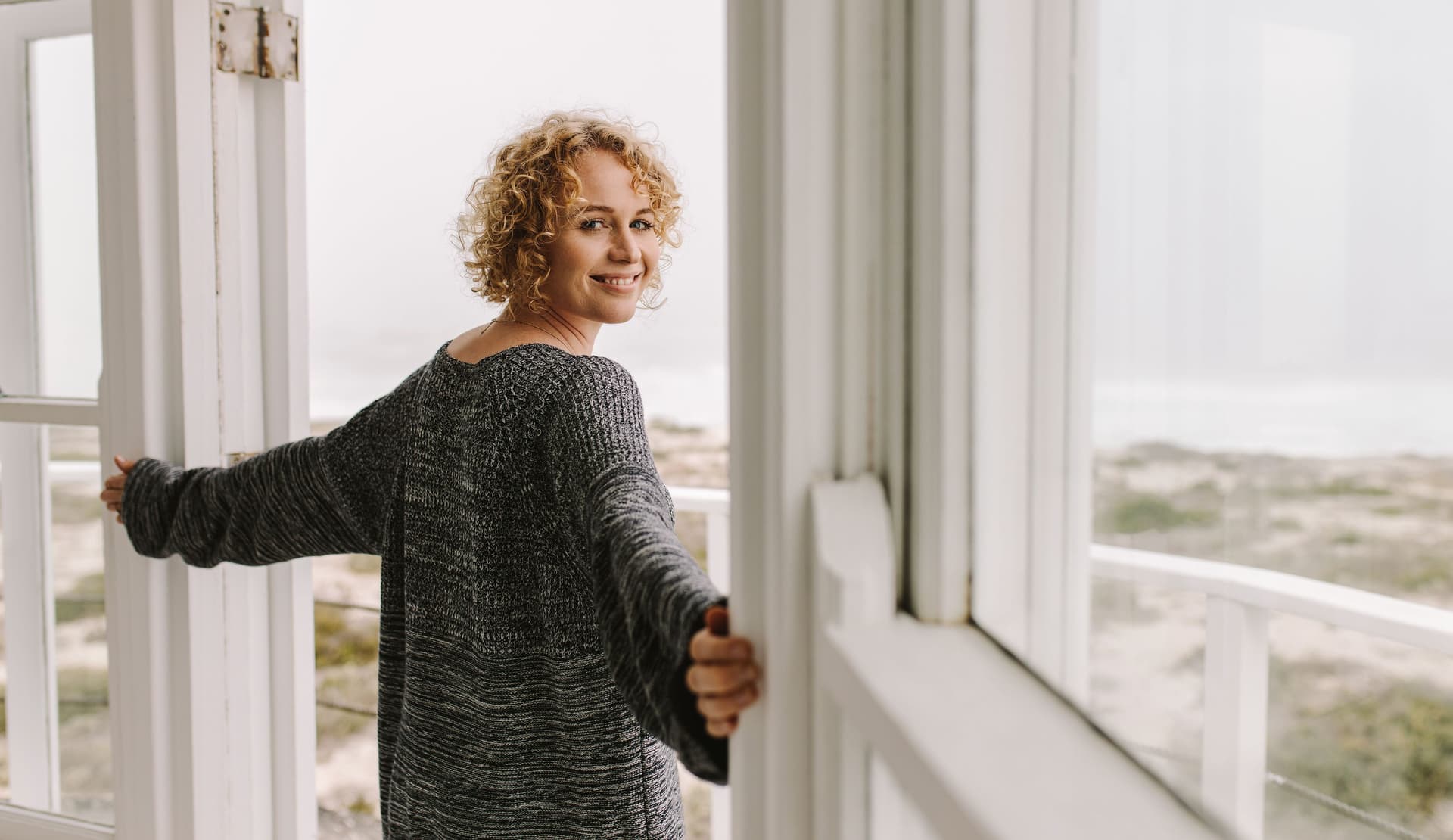 Eine Frau steht mit dem Rücken zur Kamera und macht gerade eine Terrassentür auf. Im Hintergrund ist das Meer zu sehen.