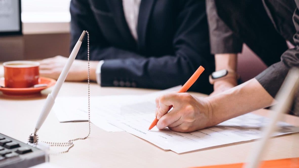 Eine Frau unterschreibt mit einem orangenen Stift ein Dokument auf einem Tisch, daneben sitzt eine weitere Person im Anzug. Auf dem Tisch liegen Unterlagen und eine Kaffeetasse. Die Szene vermittelt Vertragsabschluss oder Beratungsgespräch.