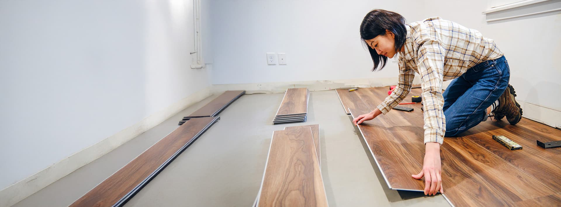 Eine junge Frau kniet auf dem Boden und verlegt sorgfältig dunkle Holzpaneele, um einen neuen Bodenbelag zu installieren. Um sie herum liegen Werkzeuge und weitere Paneele bereit.