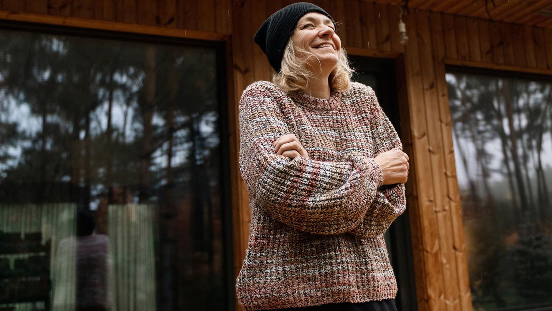 Eine Frau steht lächelnd vor einem rustikalen Holzhaus, umgeben von einer malerischen Landschaft. Sie trägt legere Kleidung und wirkt entspannt.