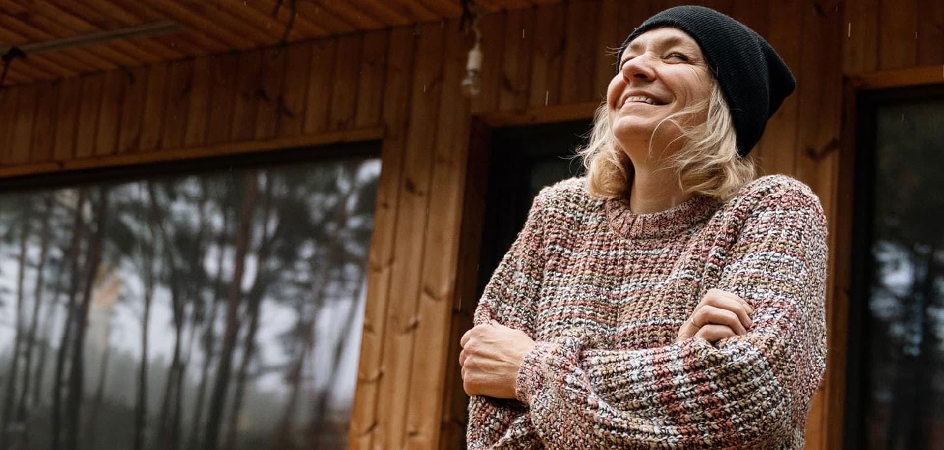 Eine Frau steht lächelnd vor einem rustikalen Holzhaus, umgeben von einer malerischen Landschaft. Sie trägt legere Kleidung und wirkt entspannt.