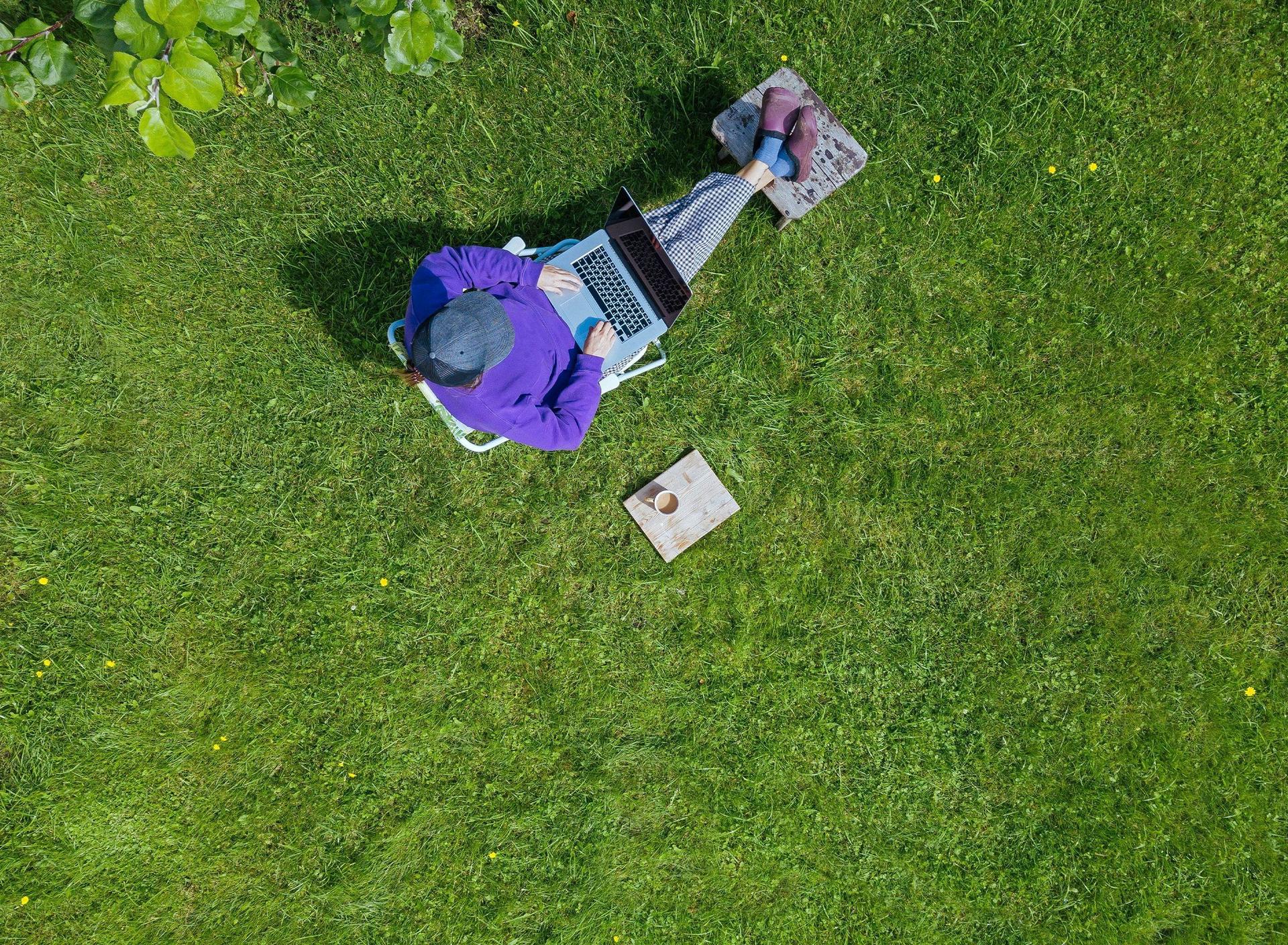 Eine junge Frau, aus Vogelperspektive fotografiert, sitzt mit ihrem Laptop auf dem Schoß und einem Kaffee neben sich bequem in ihrem Garten, auf ihrem Grundstück. 