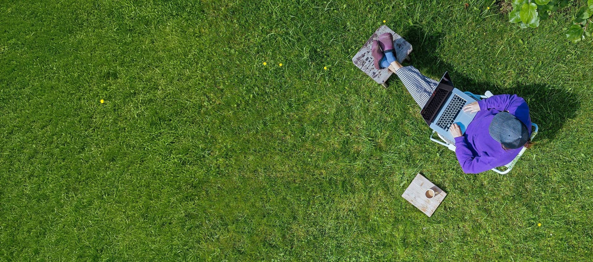 Eine junge Frau, aus Vogelperspektive fotografiert, sitzt mit ihrem Laptop auf dem Schoß und einem Kaffee neben sich bequem in ihrem Garten, auf ihrem Grundstück. 