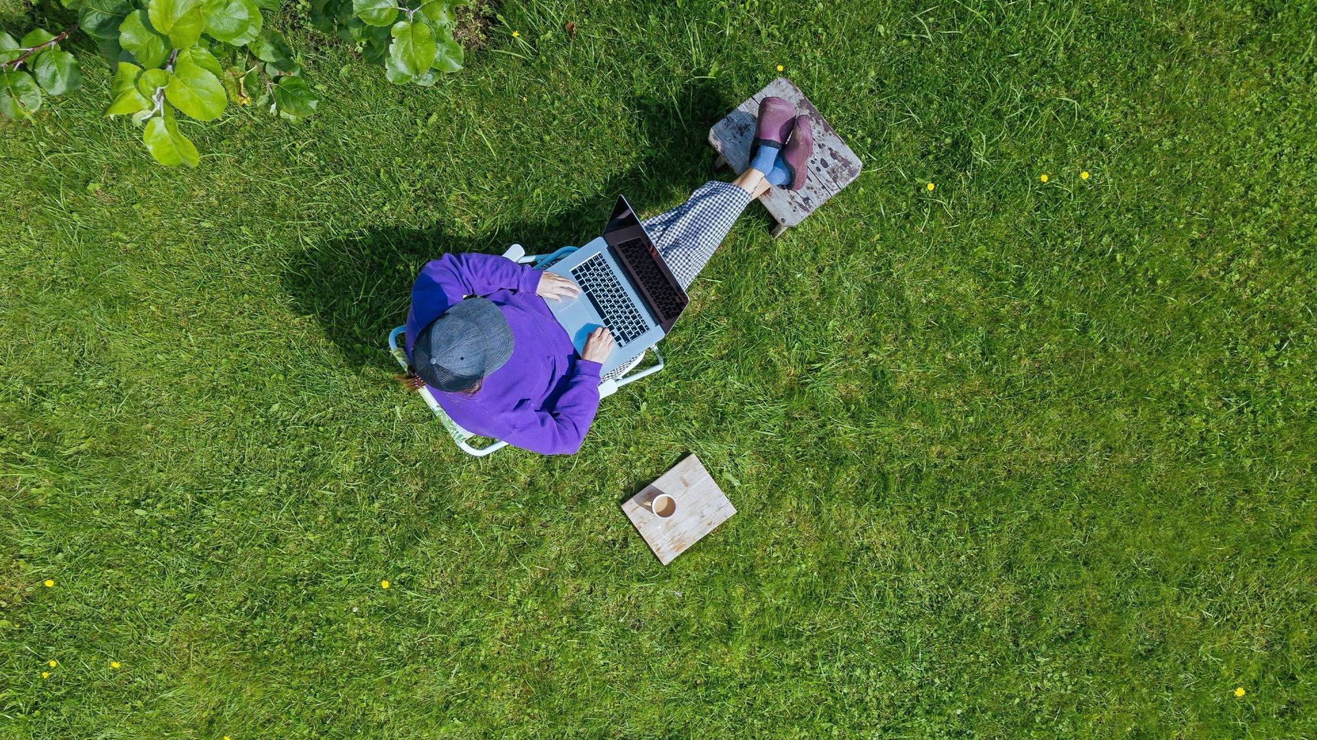 Eine junge Frau, aus Vogelperspektive fotografiert, sitzt mit ihrem Laptop auf dem Schoß und einem Kaffee neben sich bequem in ihrem Garten, auf ihrem Grundstück. 