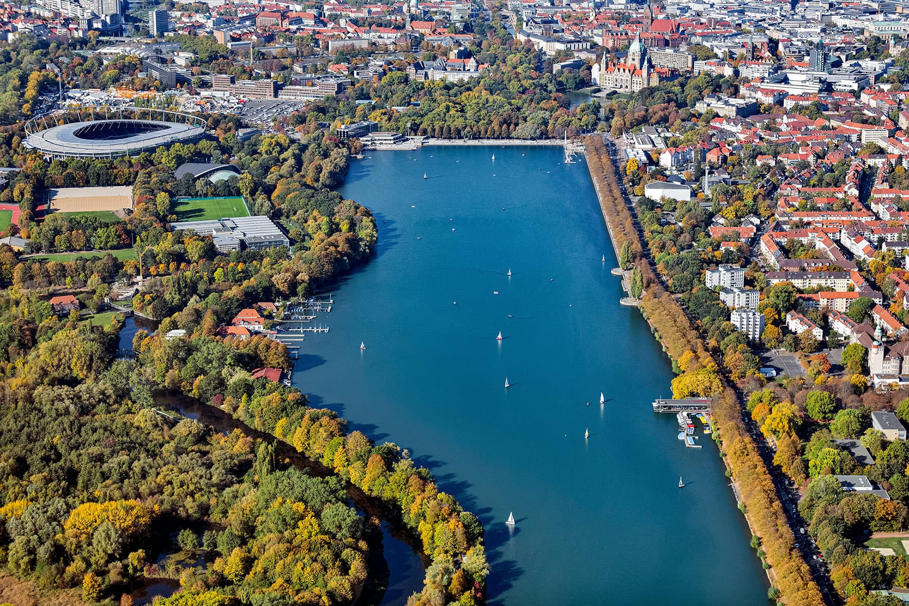 Luftaufnahme des Maschsees in Hannover mit Segelbooten, umgeben von Bäumen, Stadion und Stadtvierteln.